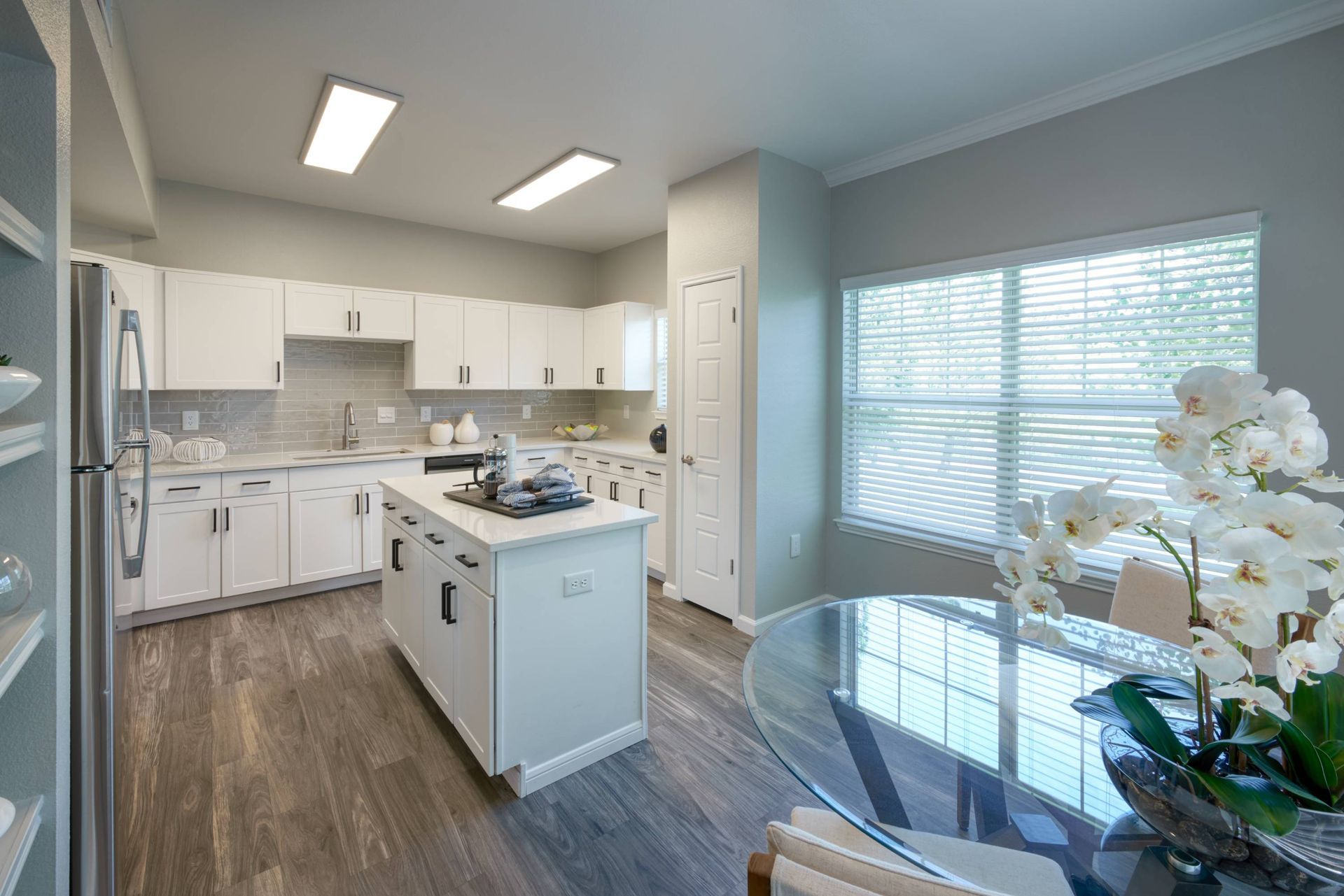 Bright white kitchen with center island, gray backsplash, and a glass dining table by a window.
