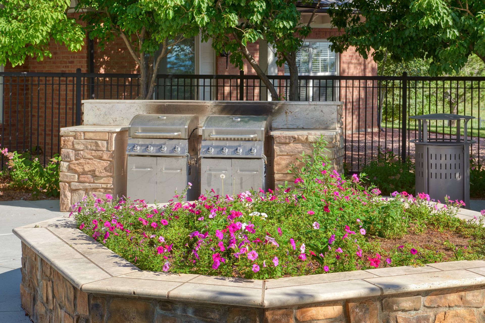 Outdoor communal grilling area with two stainless grills in a stone counter, surrounded by flowers.