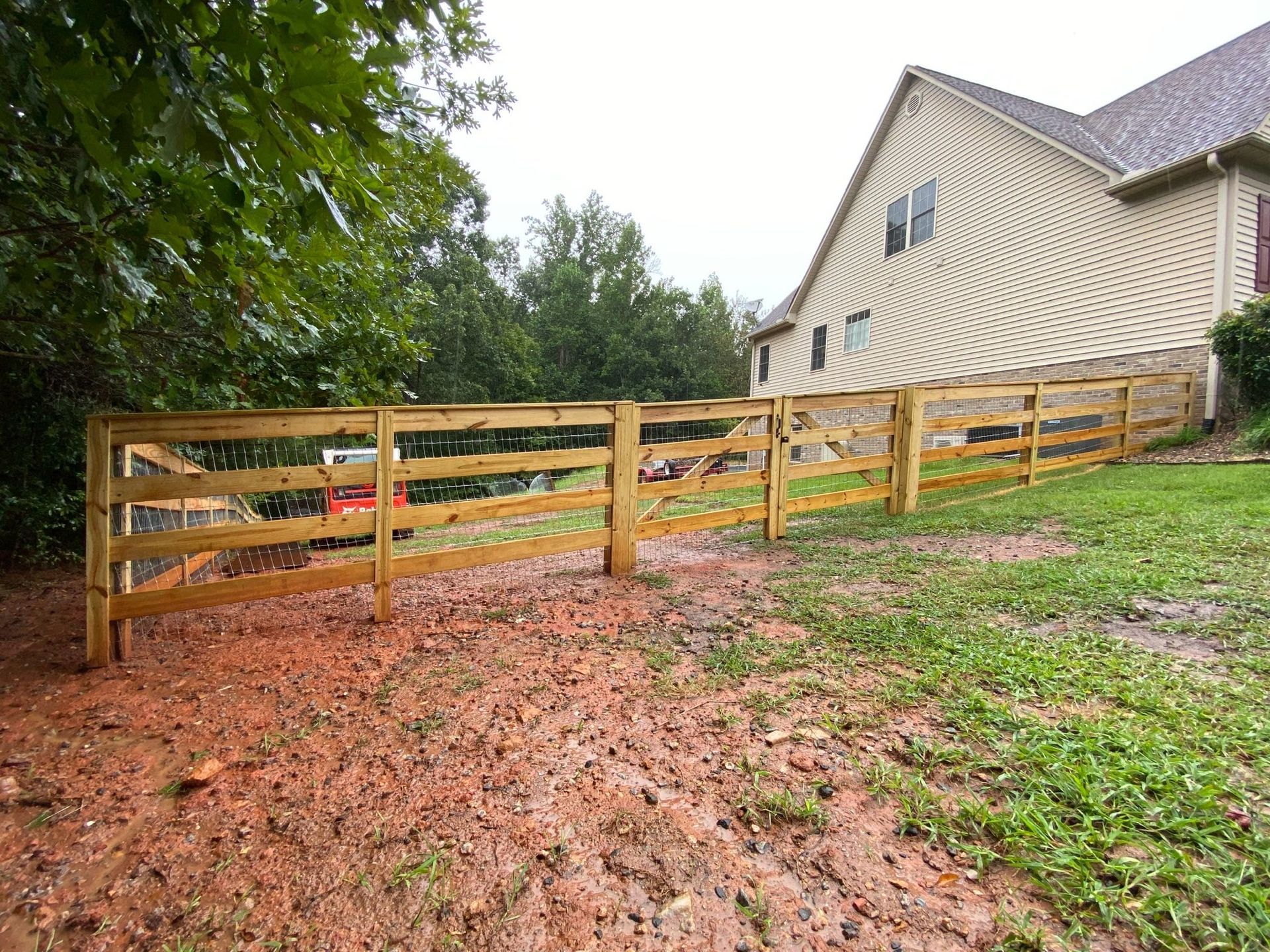 A wooden fence partially surrounds a muddy yard next to a two-story beige house and trees.