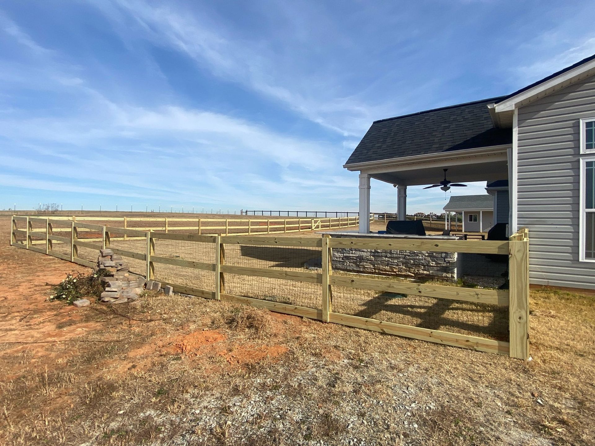 A wooden fence surrounds a house and covered patio on a sunny day. The property is on a large, open field.