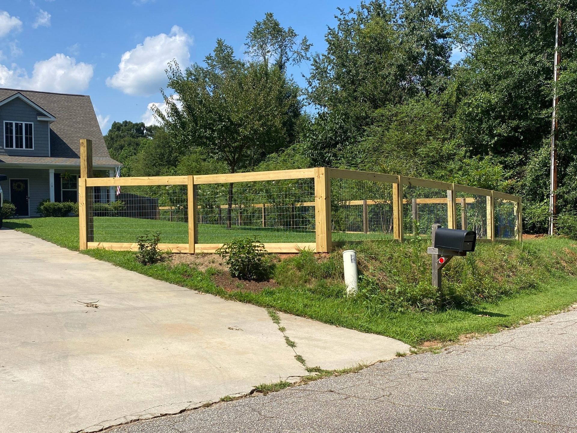 A wooden fence encloses a garden in a yard, beside a driveway and a house with blue siding. The sky is blue with clouds.