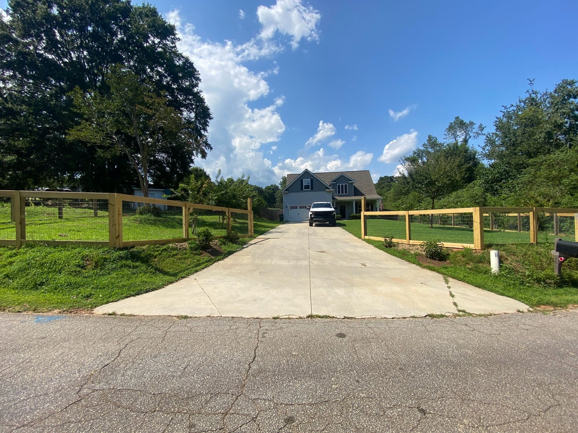 A house with a concrete driveway, a fence, and a car parked in front. The sky is blue with some clouds.