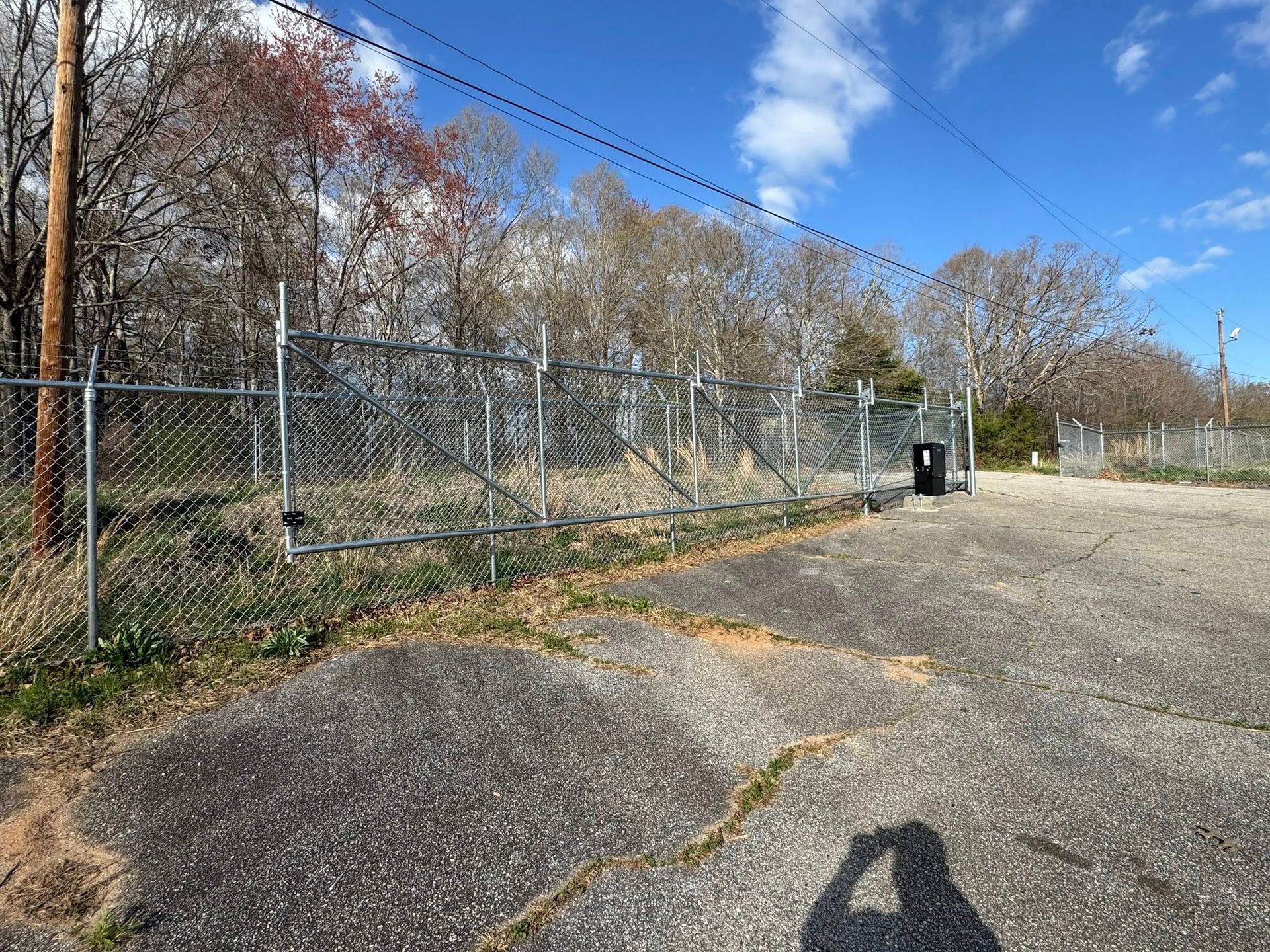 Chain-link fence with a gate blocking an overgrown area, with a gray paved lot in the foreground and trees in the background under a blue sky.