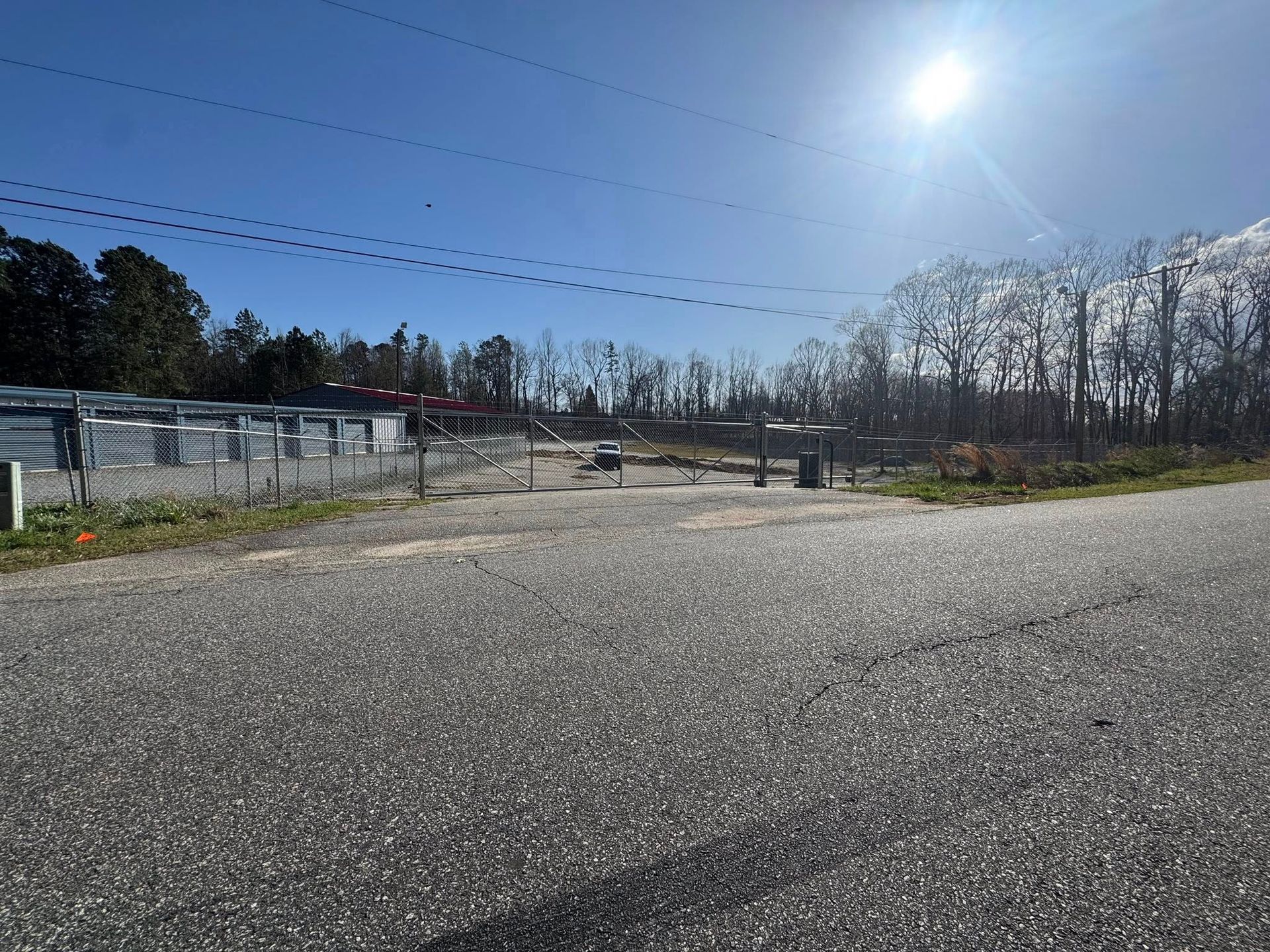 Asphalt road leads to a rural area with a fenced property, mailboxes, and trees under a bright sun.