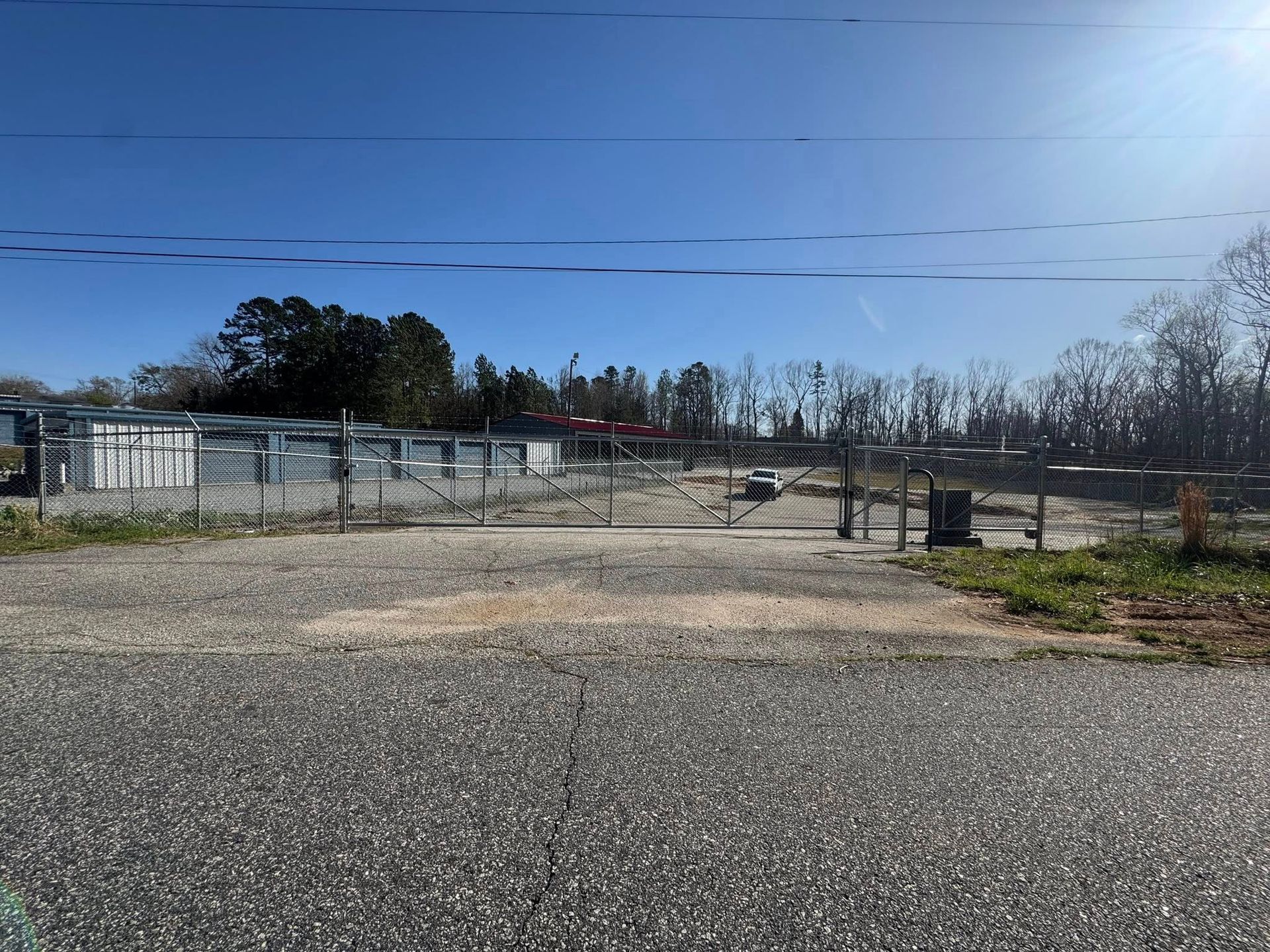 Storage units behind a chain-link fence on a sunny day. The foreground is a gravel parking area.