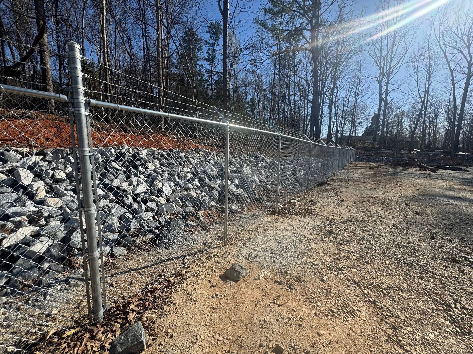Chain-link fence topped with barbed wire along a rocky embankment and dirt ground, with trees in the background under a bright sky.