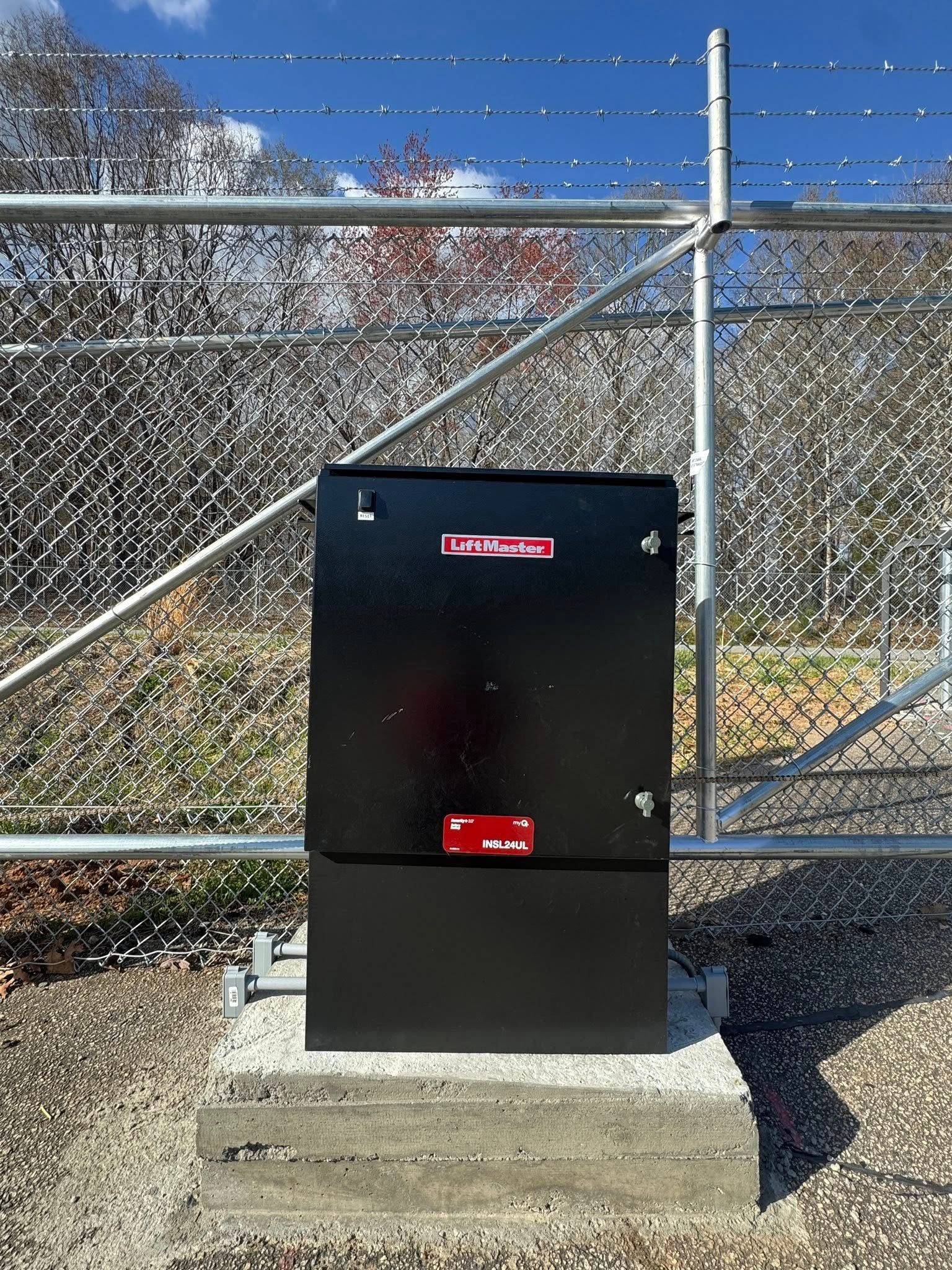 Black metal electrical box on a concrete base, in front of a chain-link fence. A red logo is visible on the box.