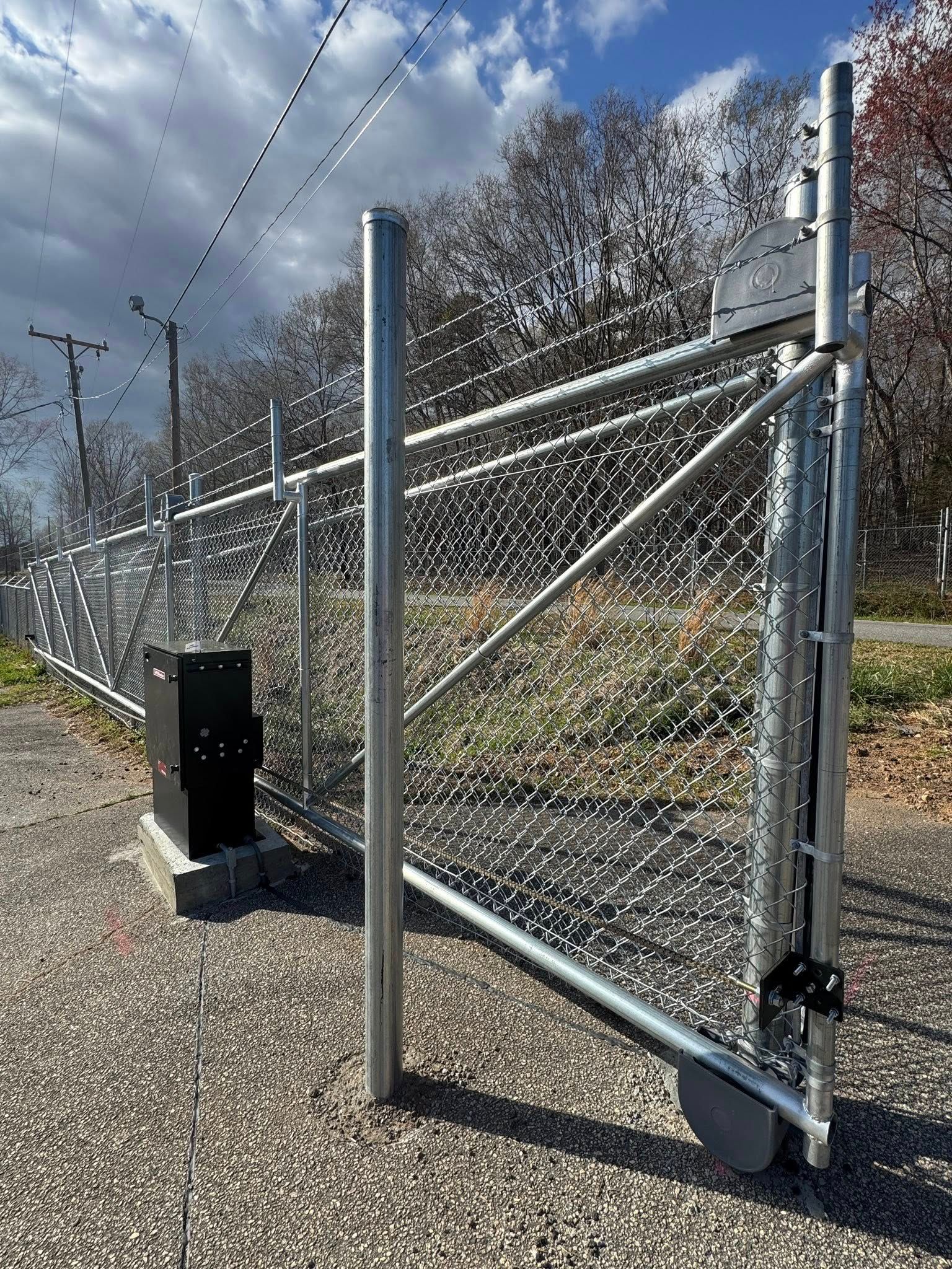 A metal gate in a chain-link fence, partially open. A black box sits next to the gate on a gravel path, with trees in the background.