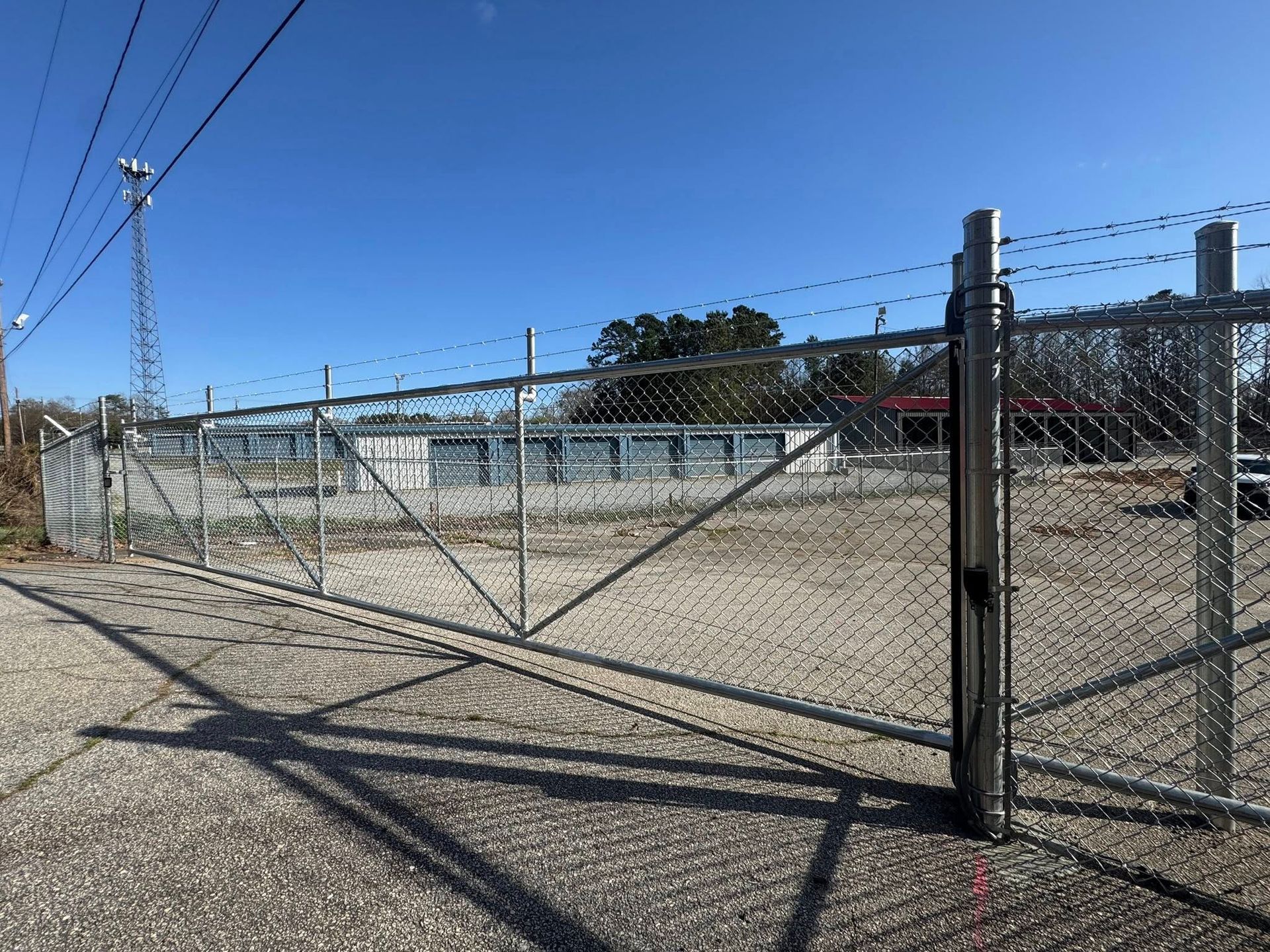 A chain-link fence with barbed wire surrounds a gravel lot with a low, gray building in the background under a blue sky.
