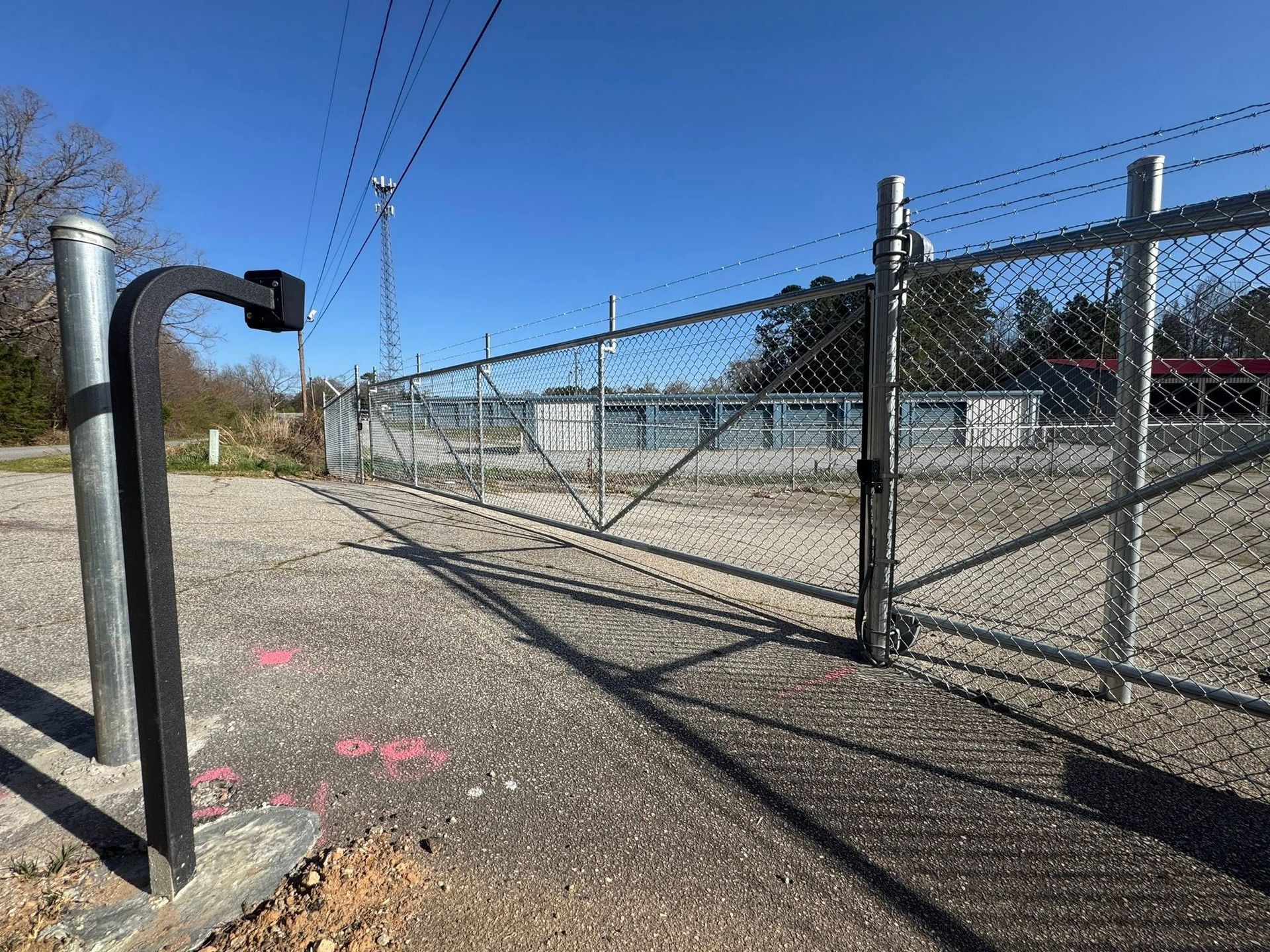 Metal gate with security arm, part of a chain-link fence, open in a gravel lot on a sunny day.