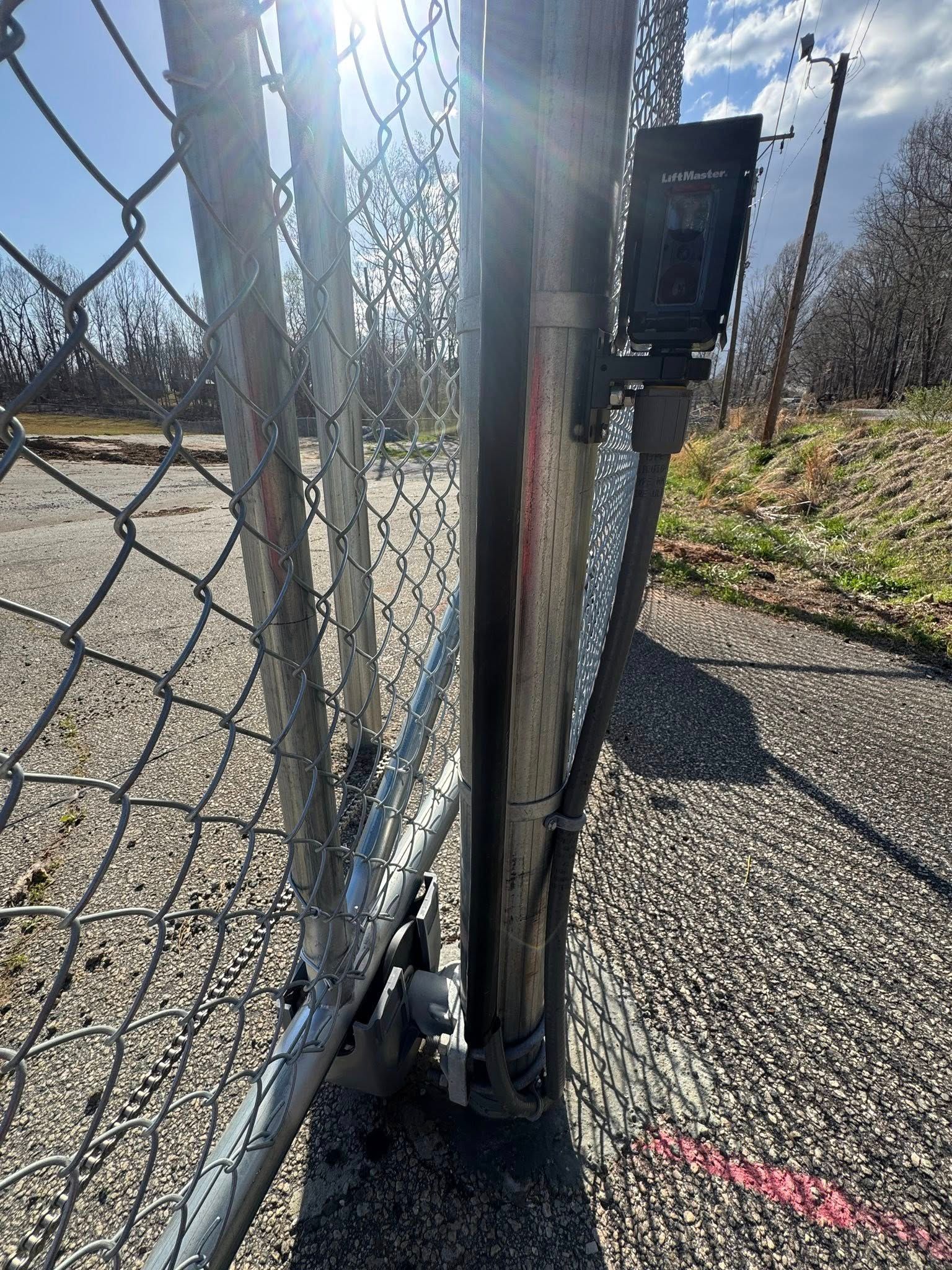 Close-up of a chain-link fence gate with security camera attached to a metal post. Bright sunlight shines from behind.