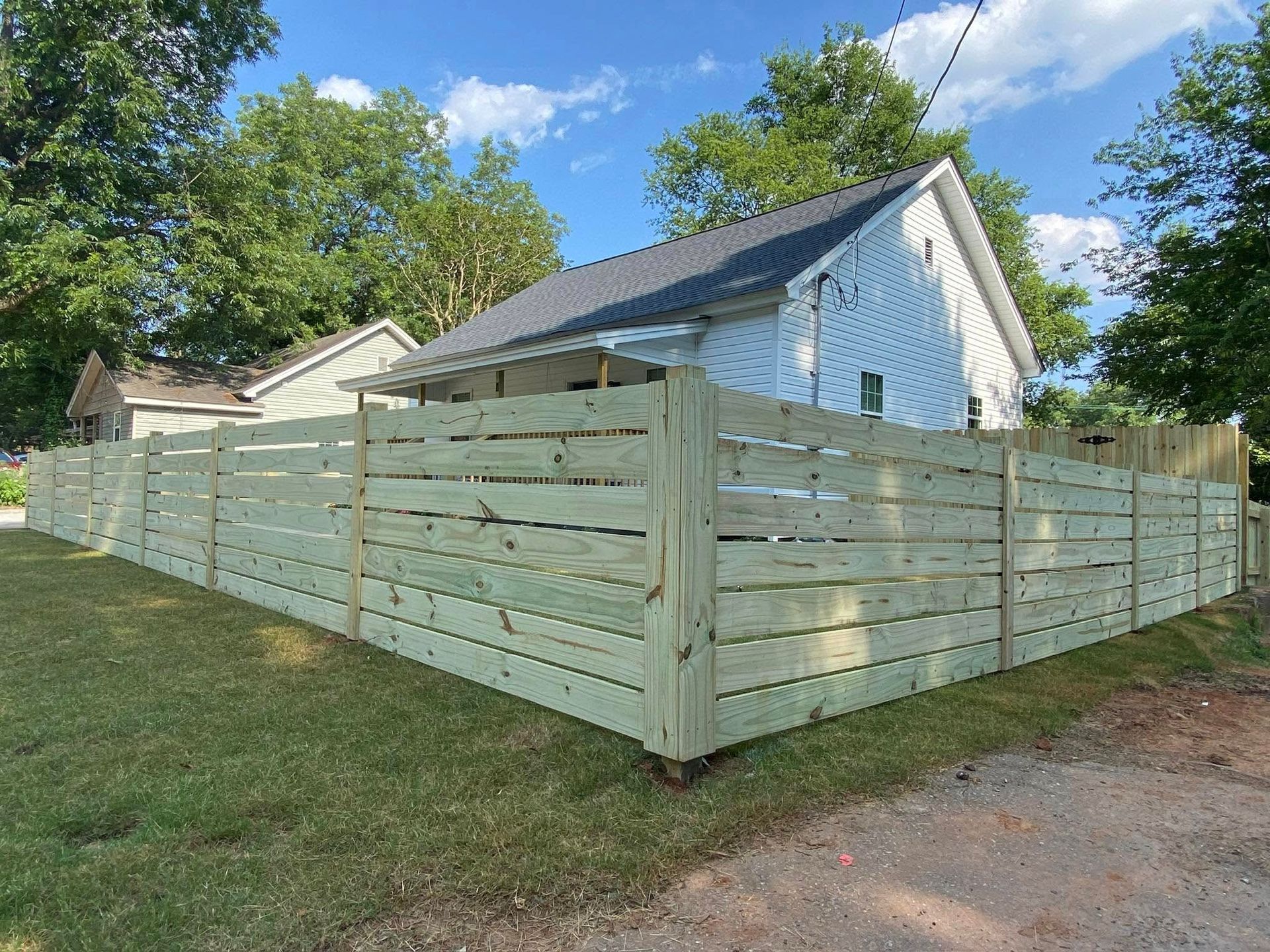 A new, horizontal-slat wooden fence surrounding a grassy yard in front of a white house under a blue sky.
