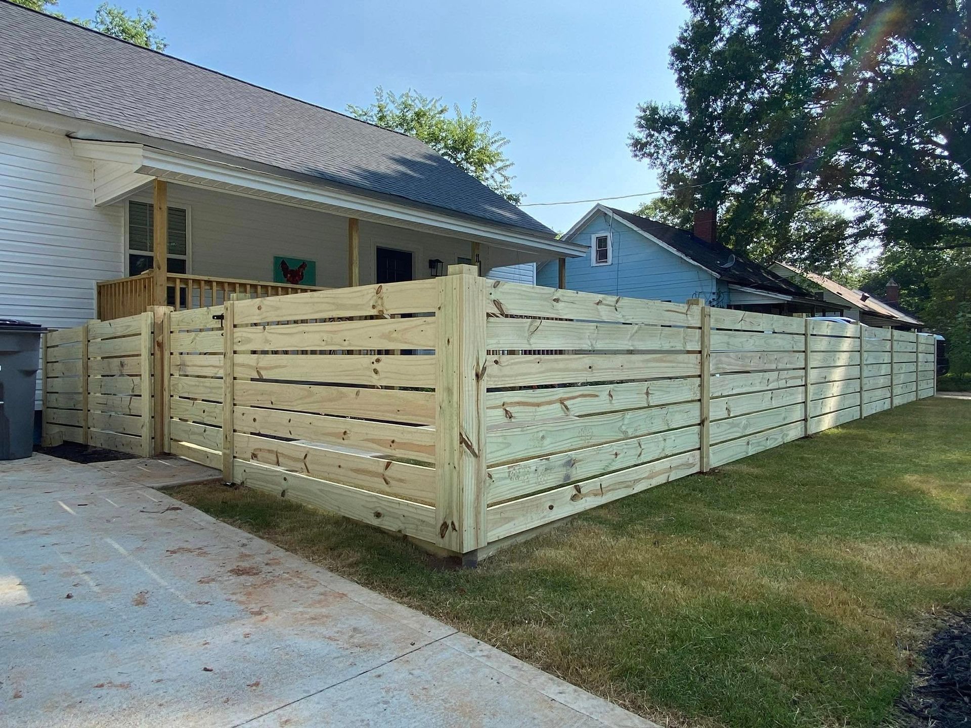 A newly-built horizontal wooden fence beside a house with green grass and a driveway. The fence is light-colored, and the house is white with a black roof.