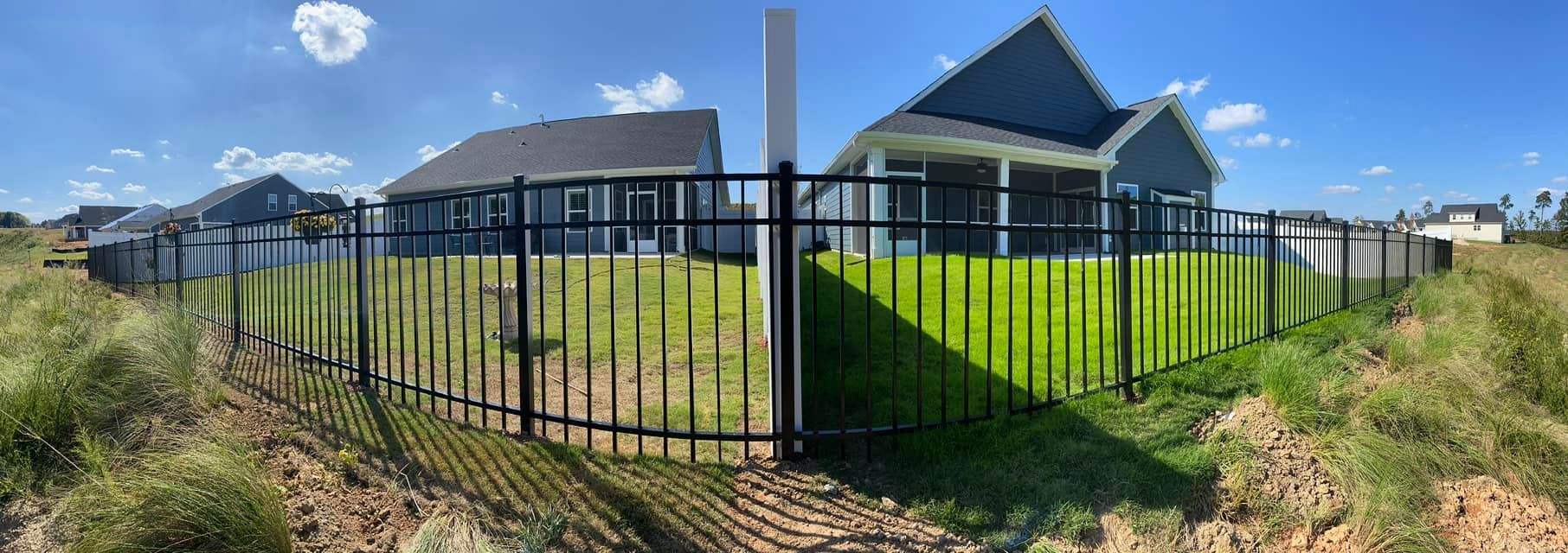 A house with a black fence and green lawn under a blue sky. Brown dirt and tall grass are on either side of the fence.