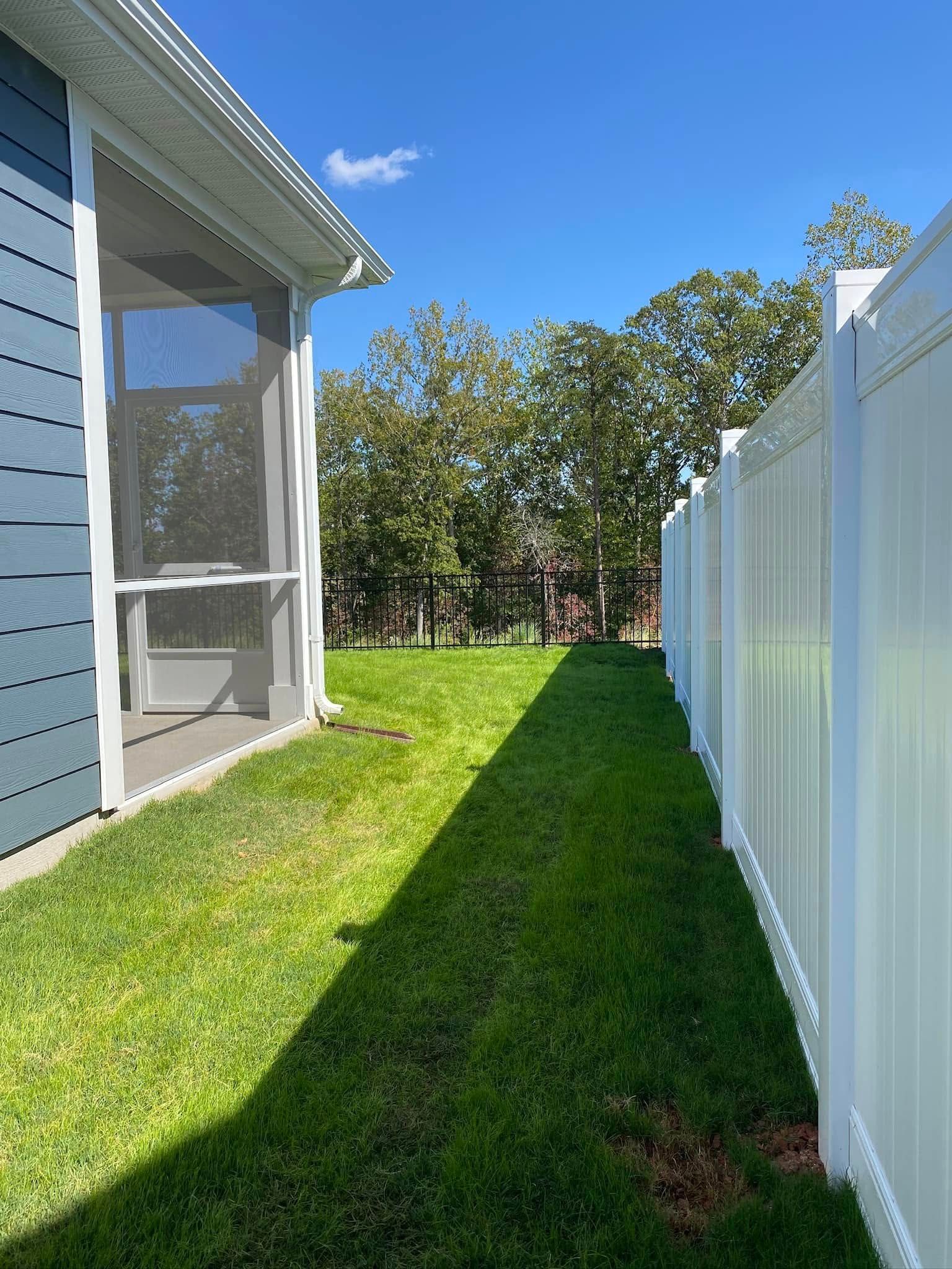 Green lawn between a blue house with a screened porch and a white vinyl fence under a bright blue sky.