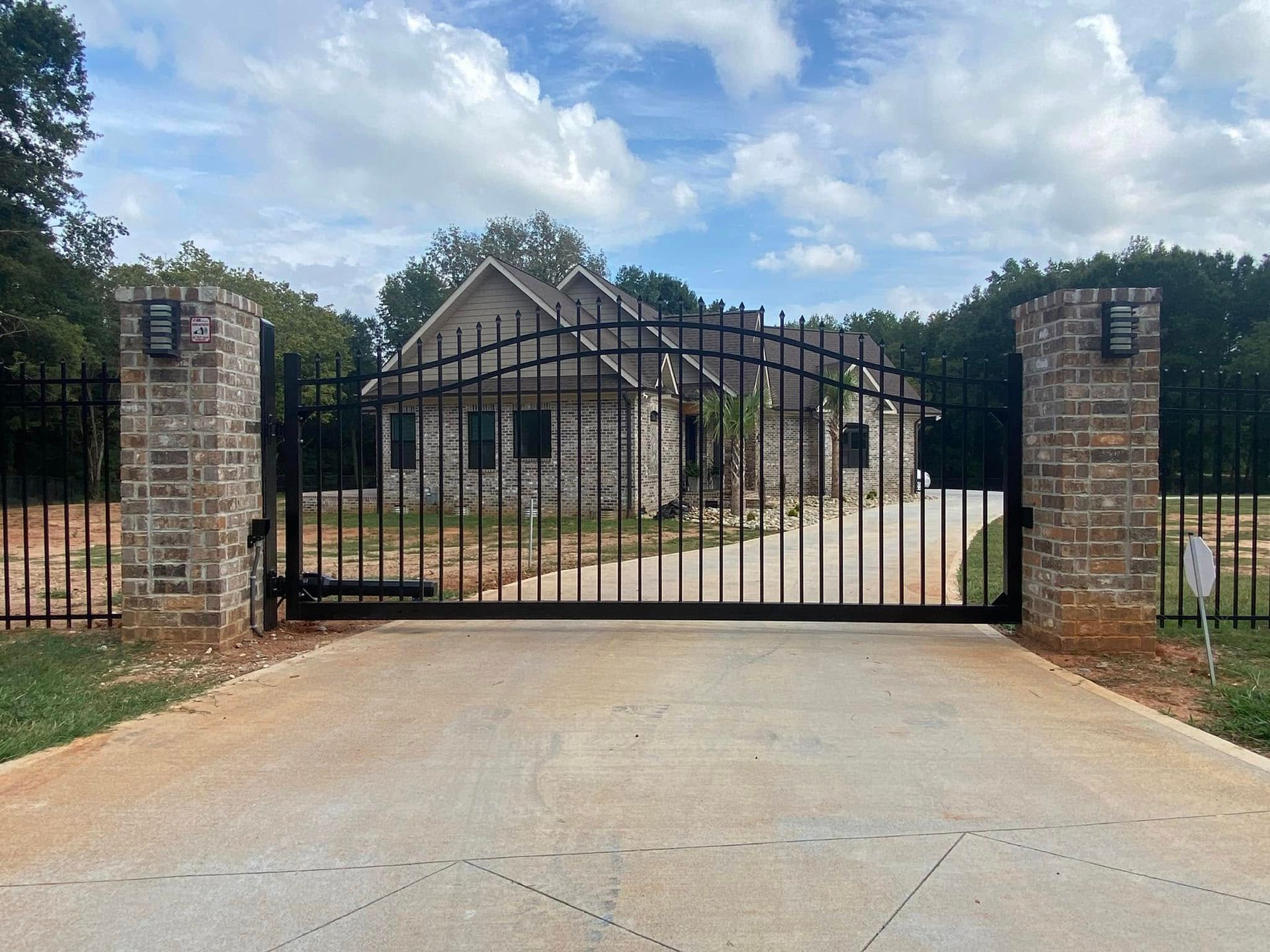 A black metal gate with brick pillars leads to a stone house on a concrete driveway under a cloudy sky.