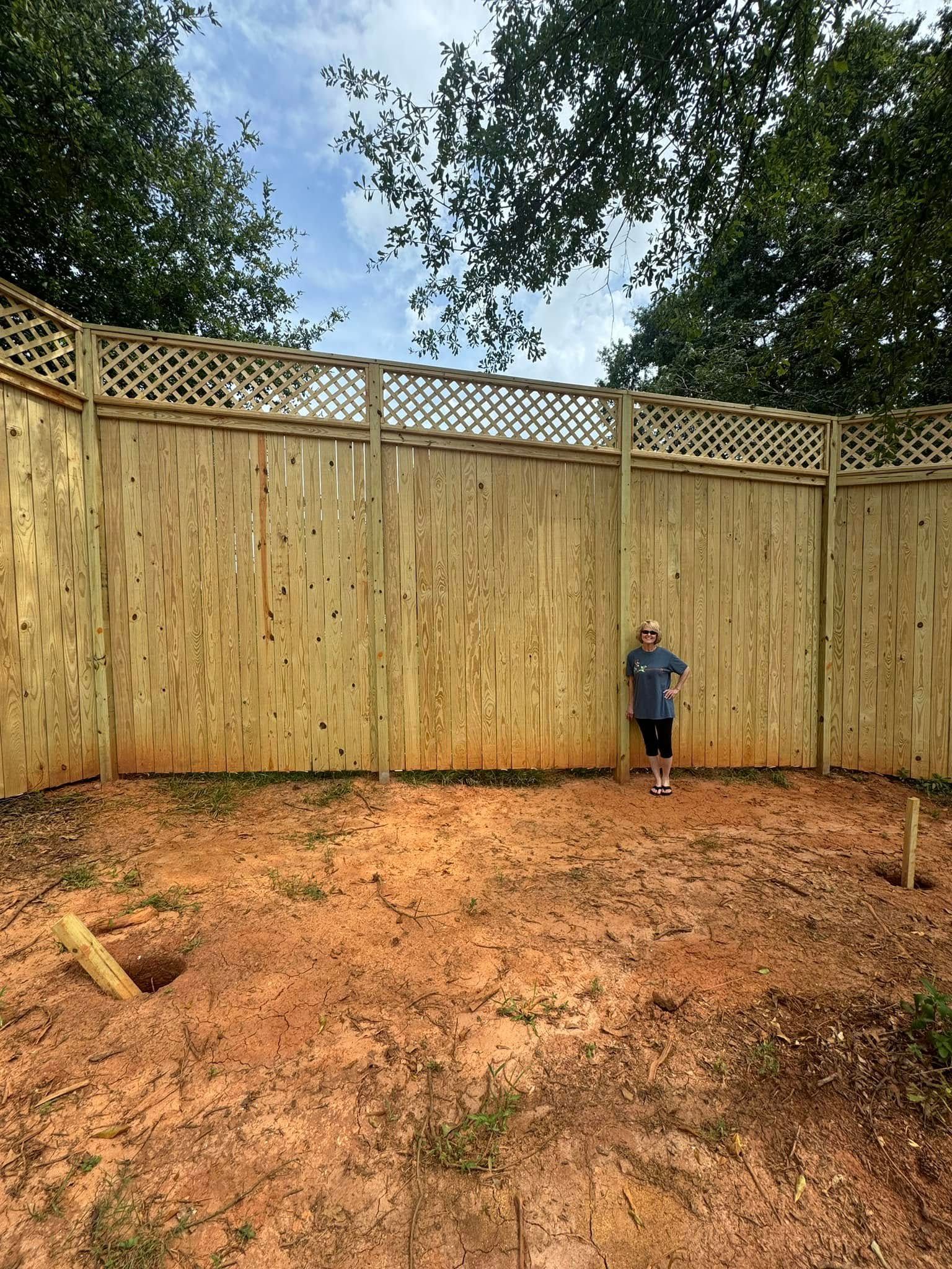 A person standing in a dirt yard enclosed by a tall wooden fence, with trees and a cloudy sky in the background.