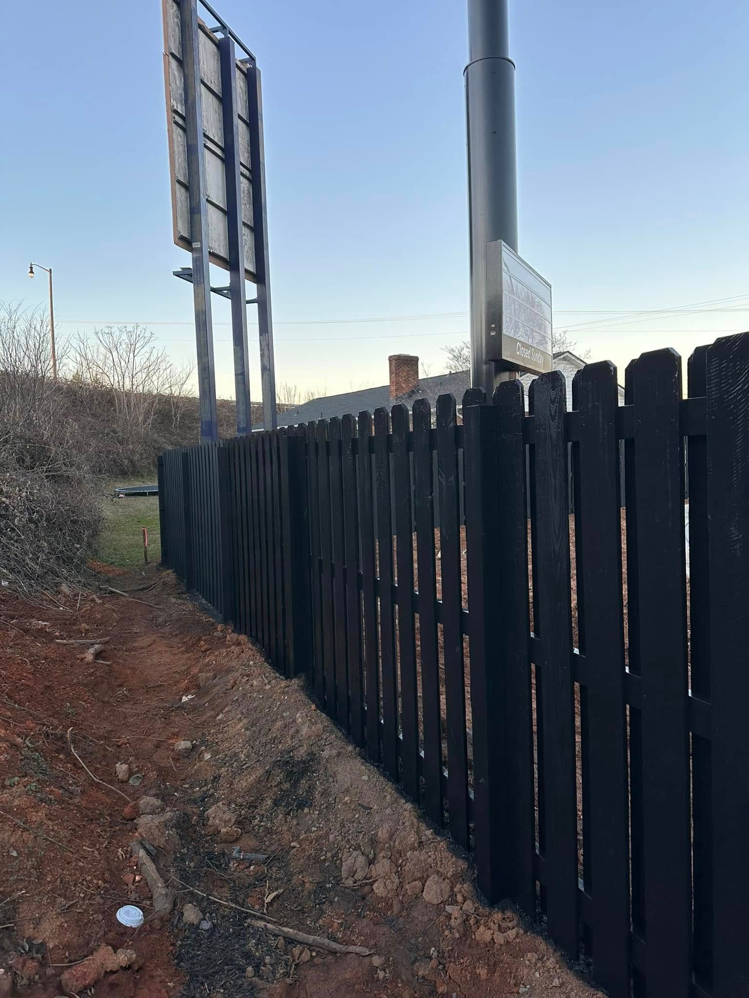 Black wooden fence runs along a dirt edge, next to a tall pole with signs, under a clear sky.