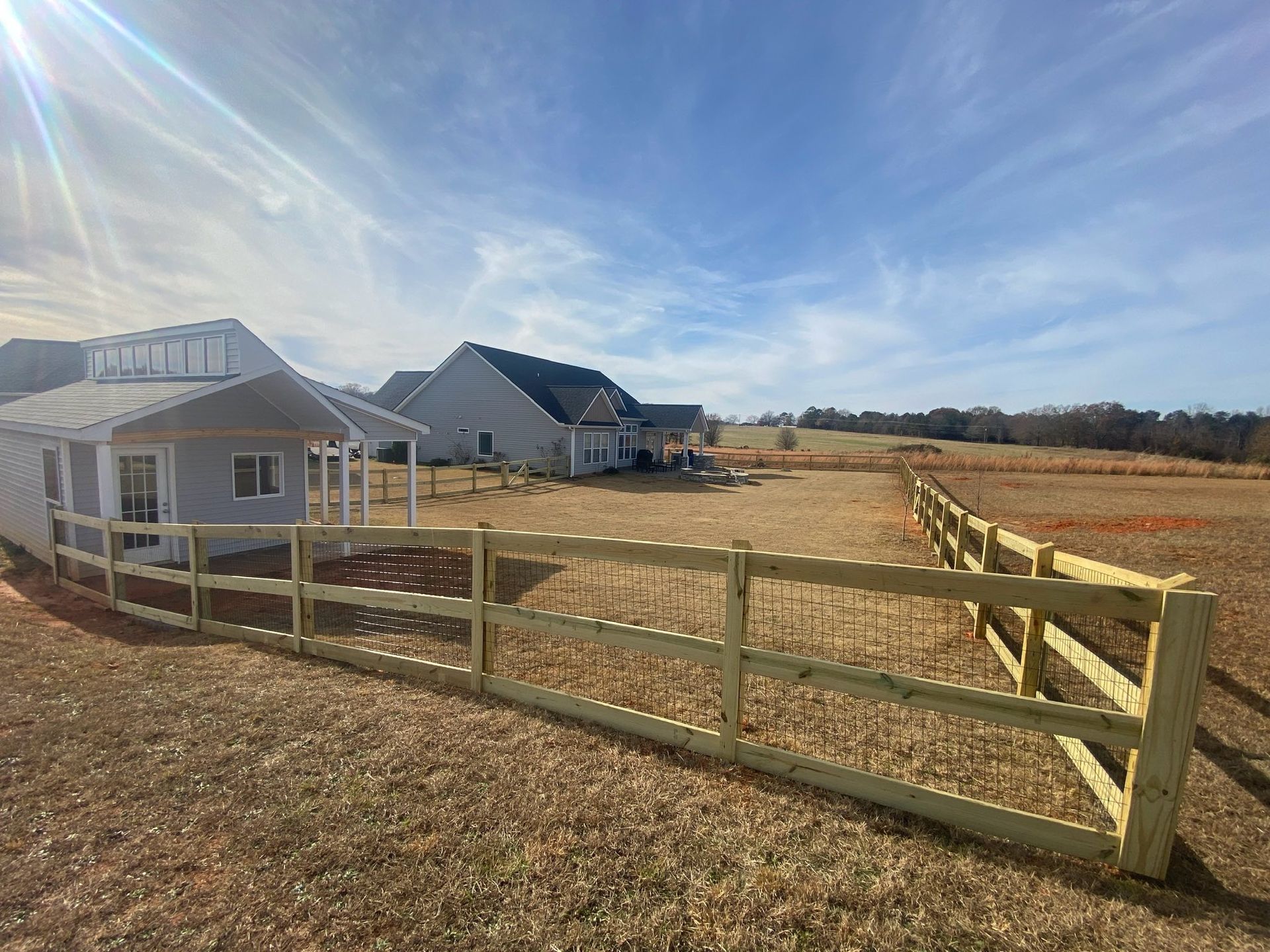 Wooden fence in a field with two houses in the background under a blue sky.