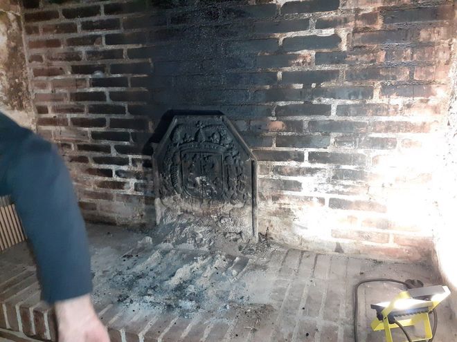Cheminée en briques avec des cendres et de la suie, une grille à feu en métal et une lampe torche jaune sur le foyer ; une personne est partiellement visible à gauche.