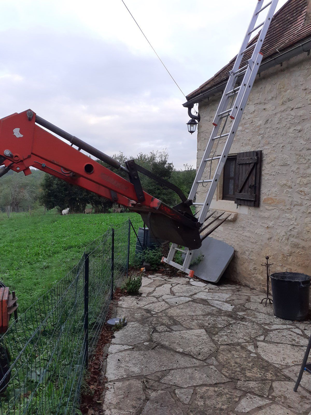 Un chariot télescopique rouge soulève une échelle contre le toit en pierre d'une maison près d'un jardin.
