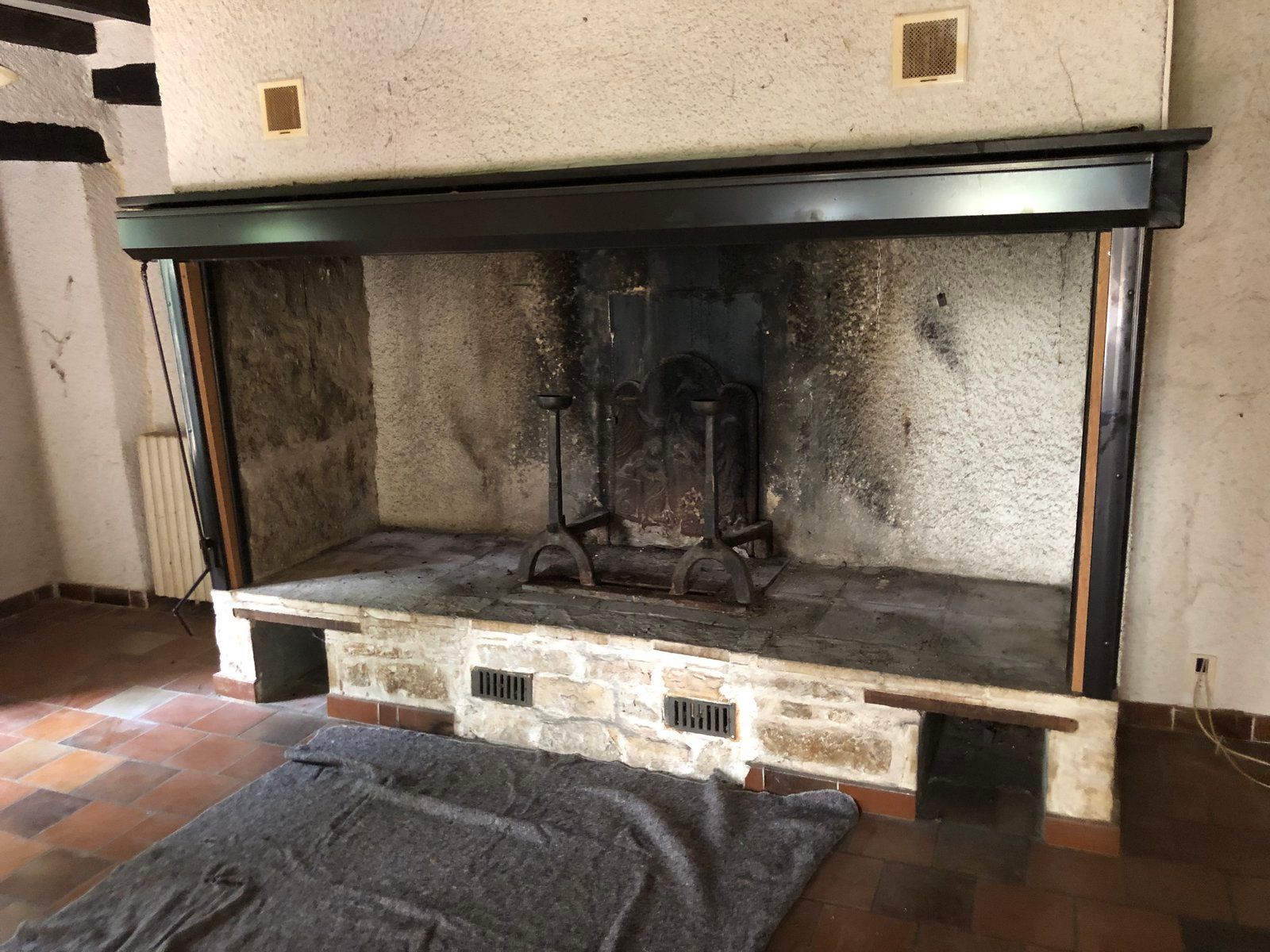 Cheminée en pierre avec manteau noir et foyer noirci par la suie dans une pièce carrelée