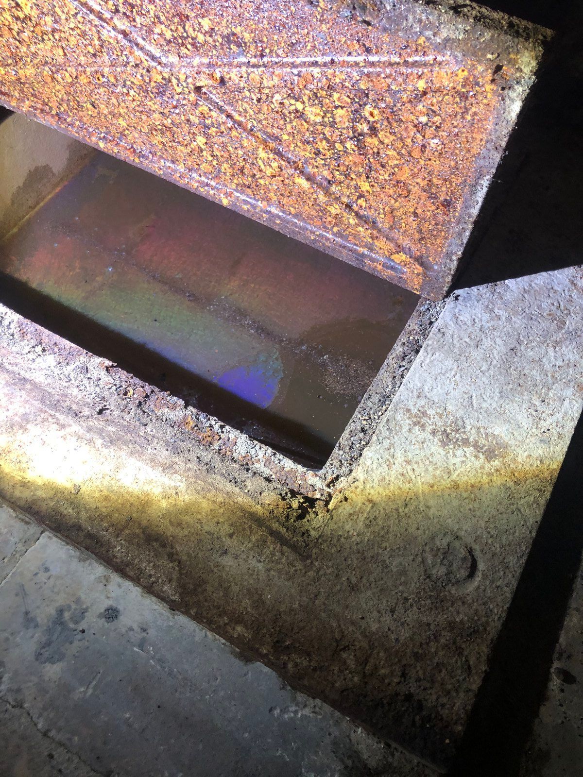 Un regard ou un drain rectangulaire ouvert et humide, entouré de béton et de métal rouillé.