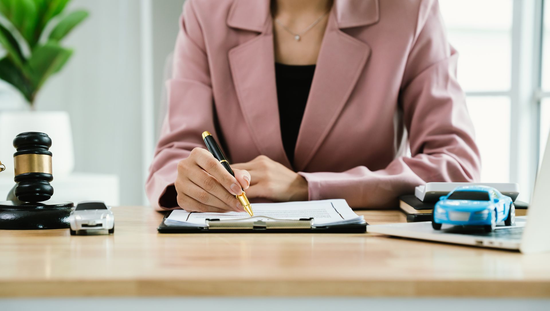 Lawyer writing on legal documents at a desk with a gavel and toy car models nearby.