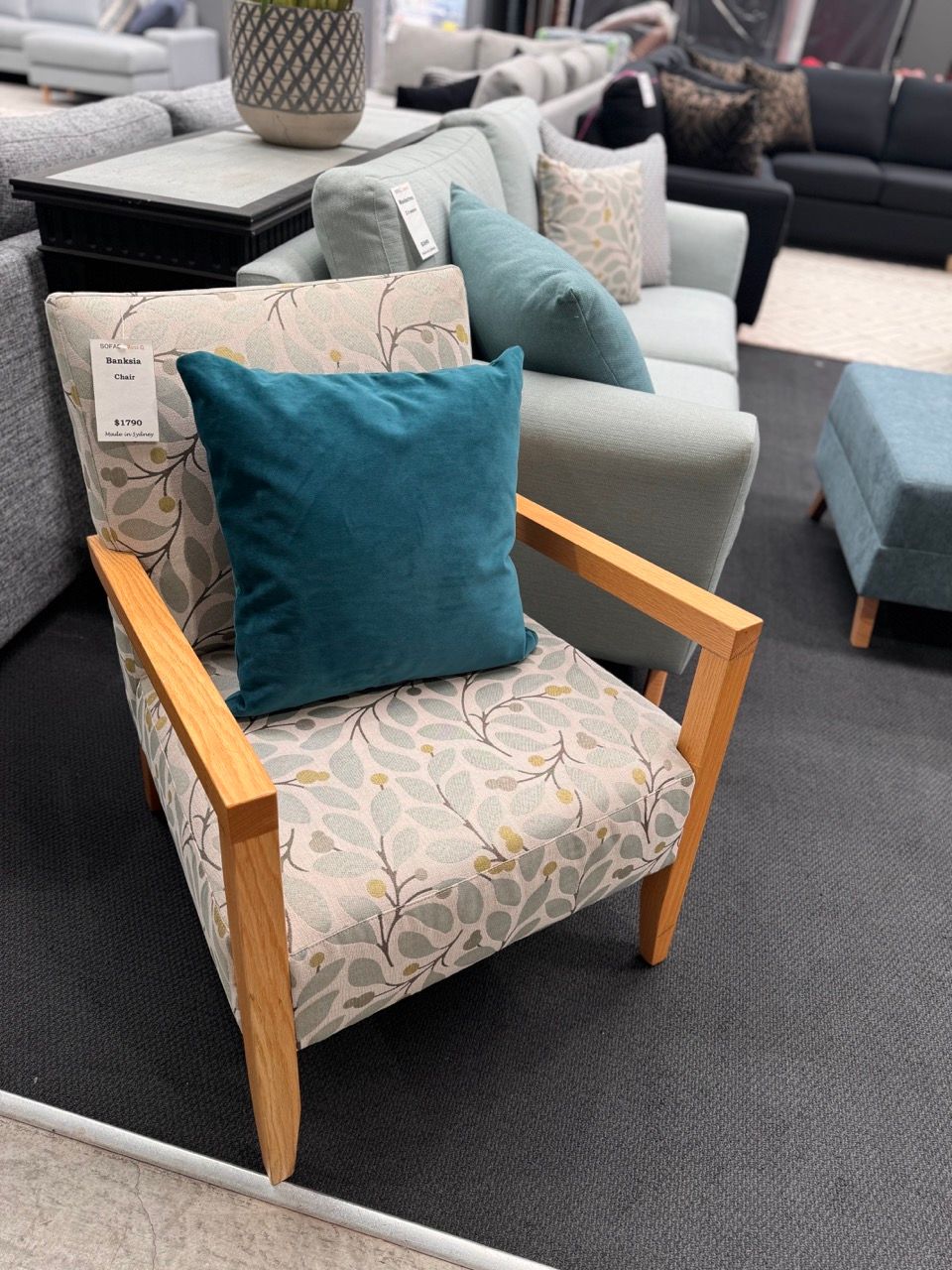 Armchair with light wood frame and patterned fabric, teal pillow, in furniture store.