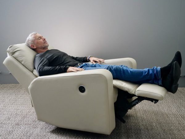 Man reclined in a cream-colored chair, eyes closed, in a gray-walled room.