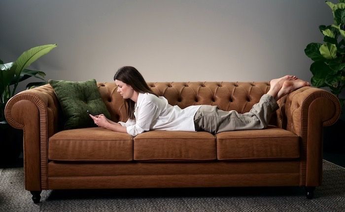 Woman lying on brown couch, looking at her phone; green pillow, plants.