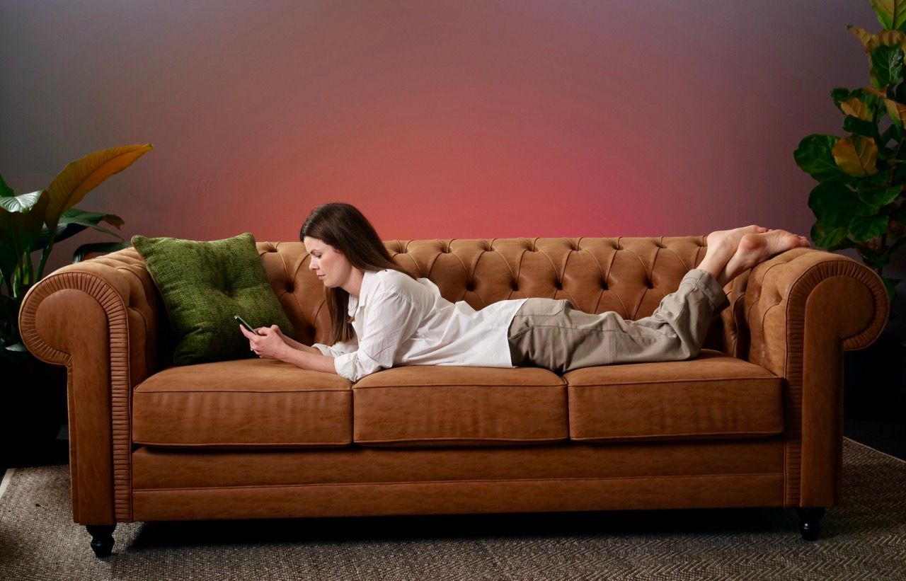 Woman on a brown couch, looking at her phone. Green pillow, pink and purple background.