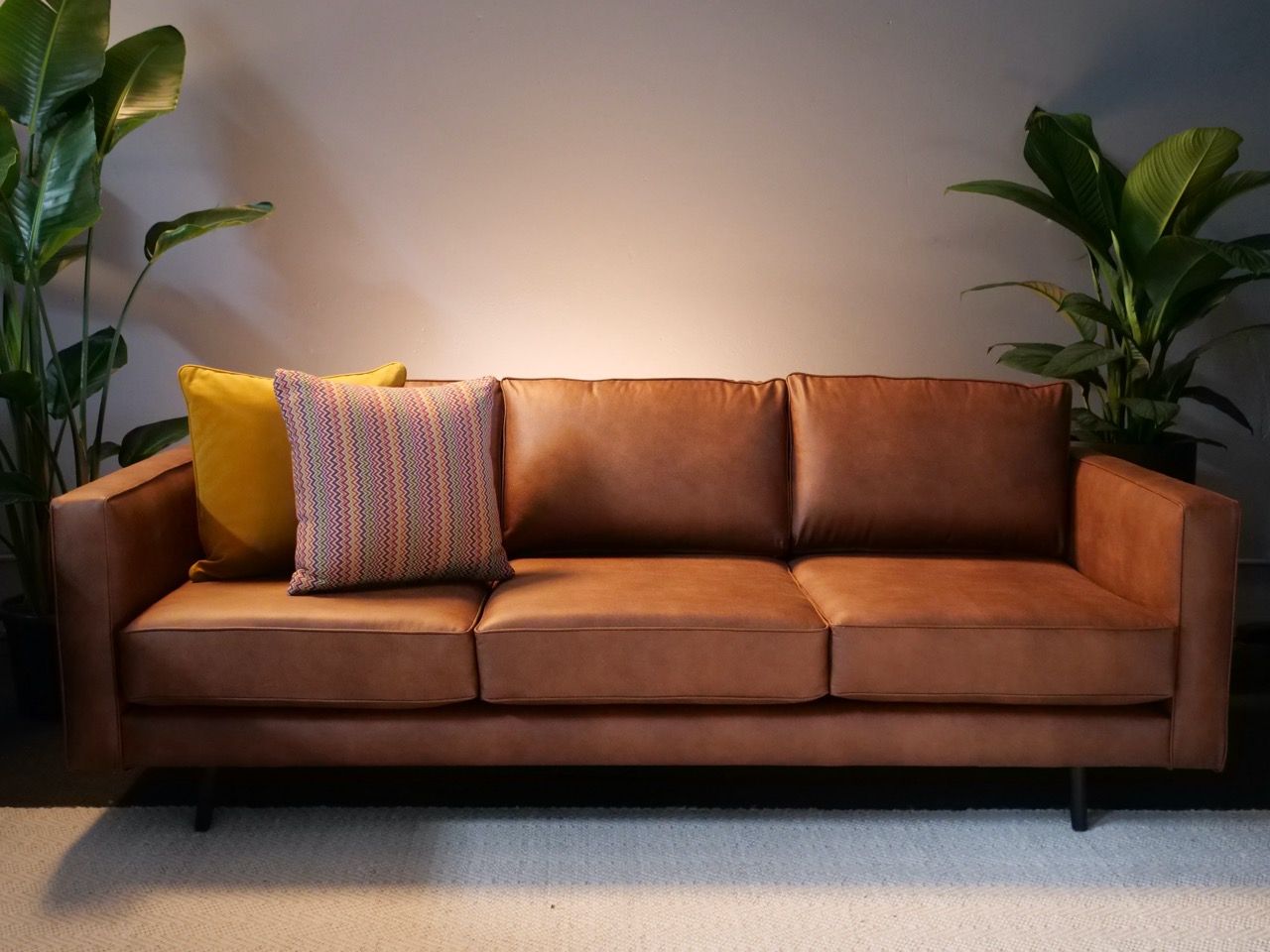 Brown leather sofa with yellow and patterned pillows, flanked by potted plants.
