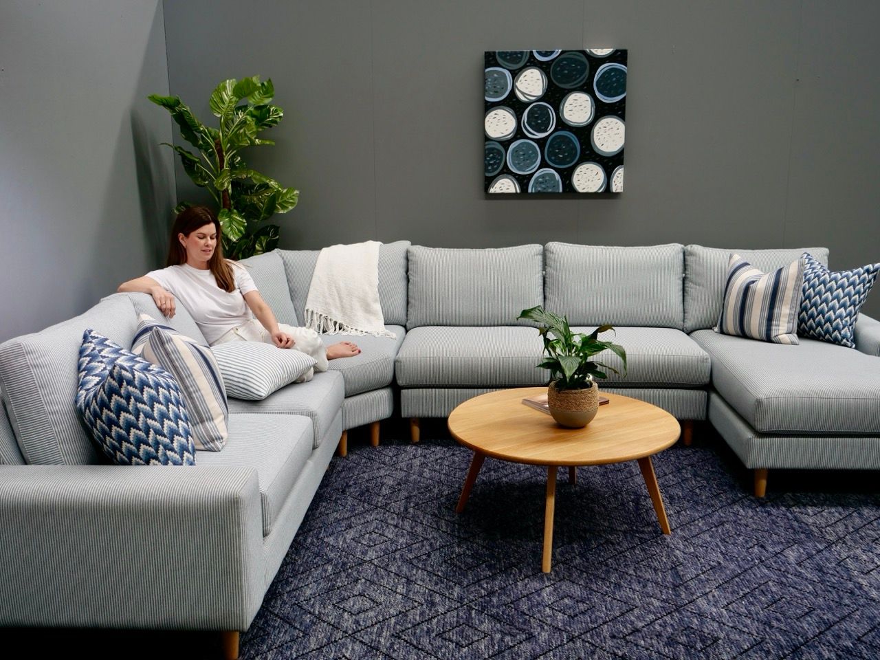 Woman relaxing on a light blue sectional sofa in a gray-walled room with a wooden coffee table and artwork.