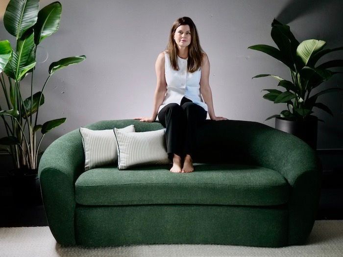Woman sitting barefoot on a green couch, flanked by potted plants, in a modern room.