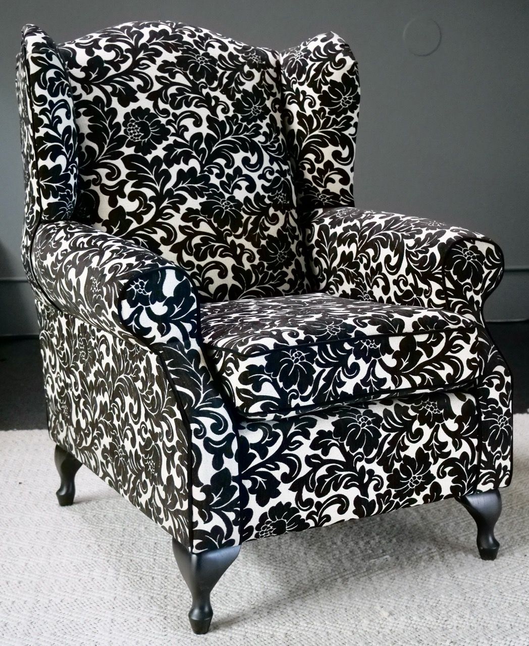 Wingback chair upholstered in black and white floral fabric, on a rug, against a gray wall.