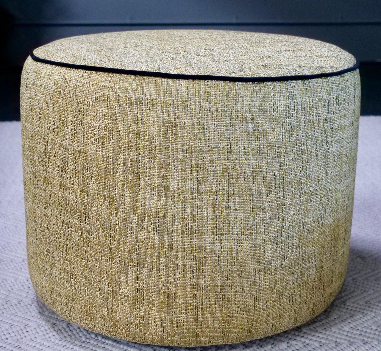 Tan upholstered cylindrical ottoman with black piping.