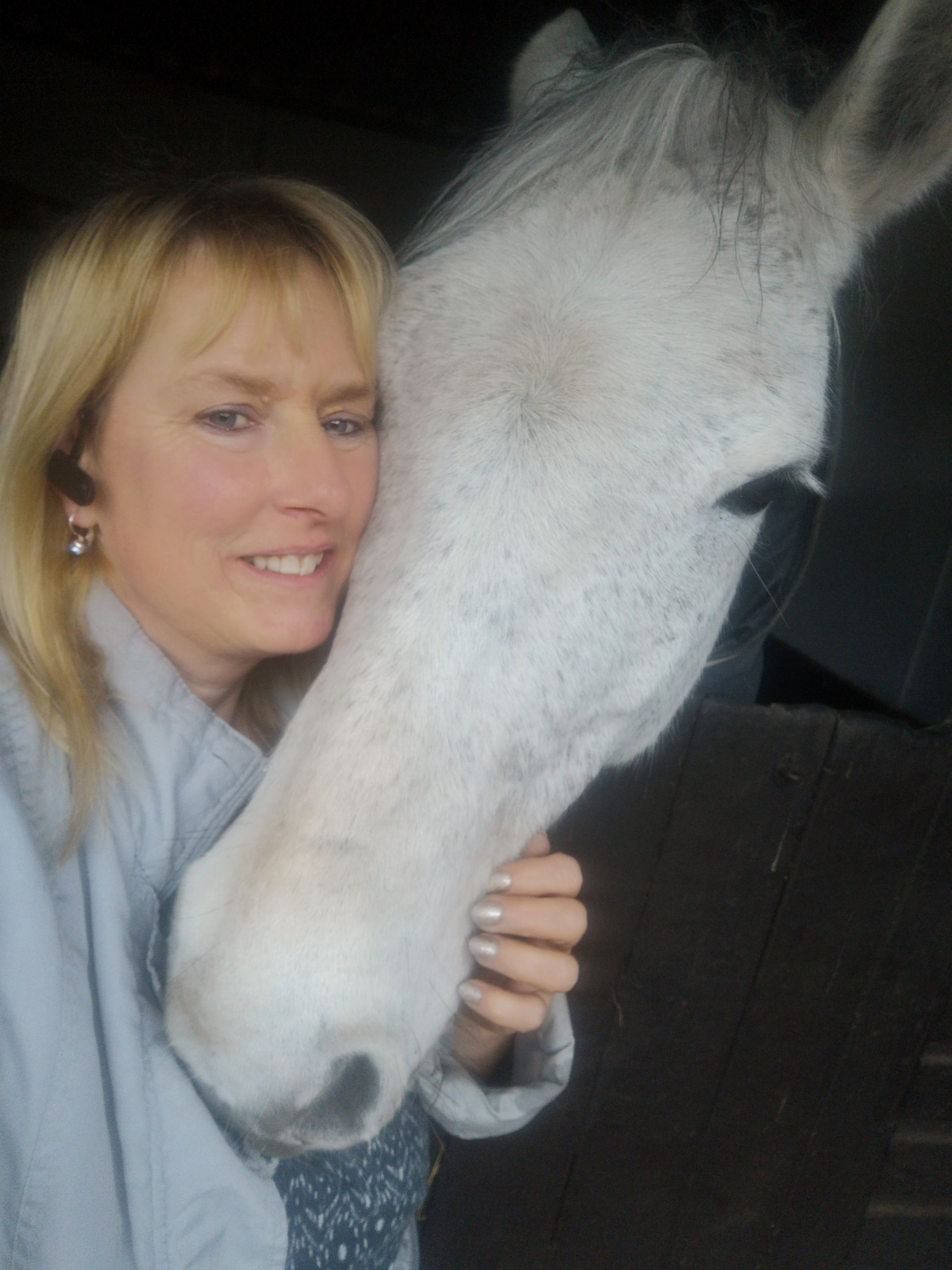 A mature woman smiles as a white horse nuzzles into her with their nose.