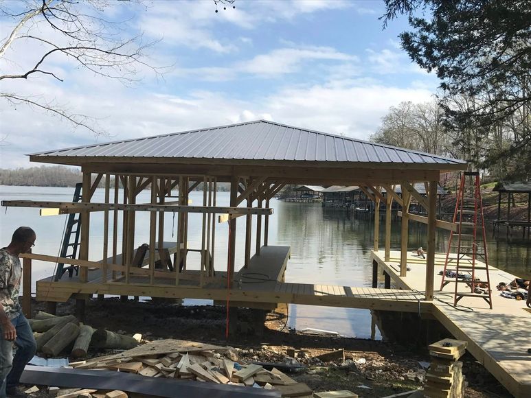 A wood-framed boat dock under construction by a lake, featuring a metal roof and a construction worker standing nearby.