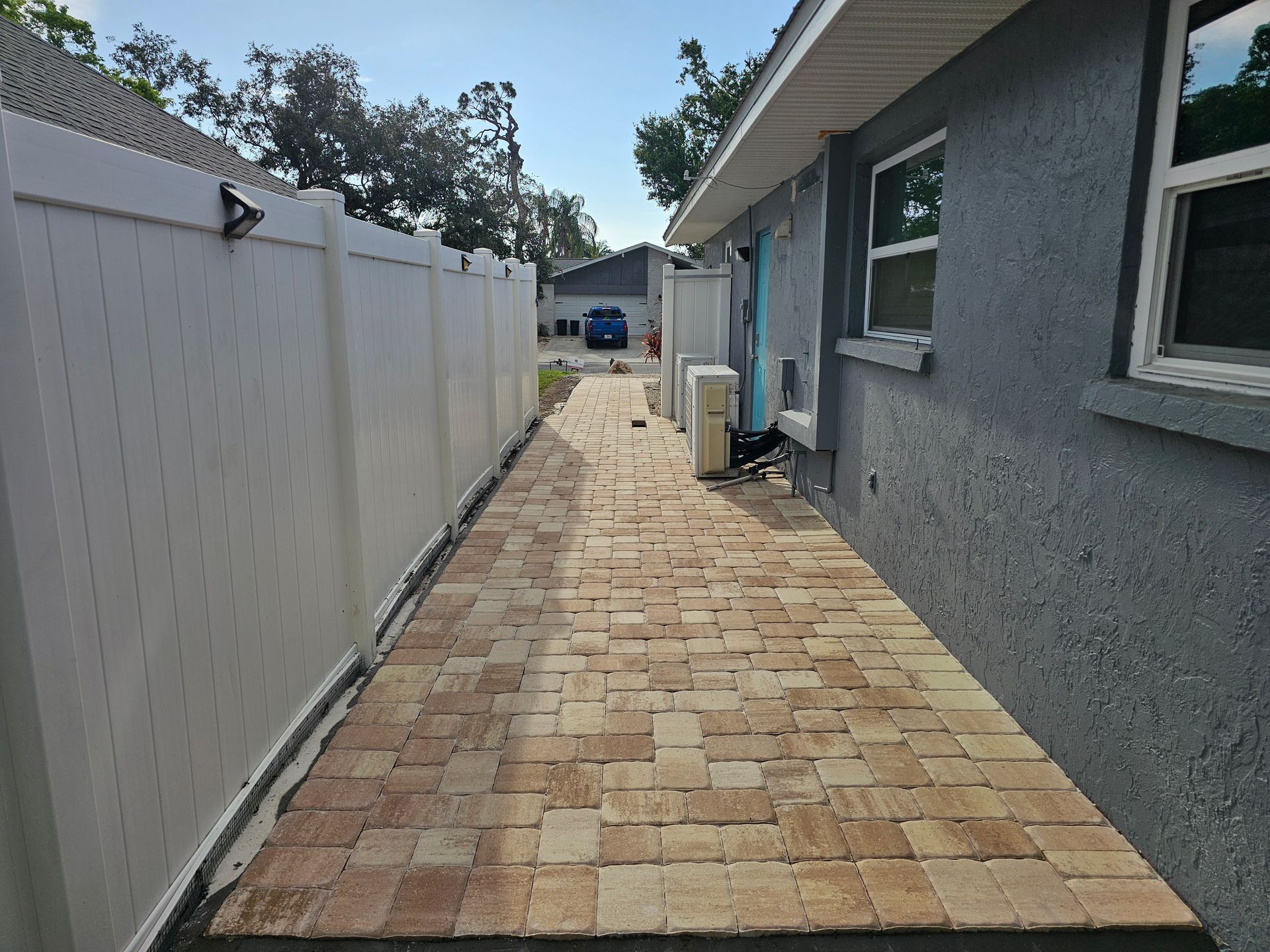 Brick walkway between a white fence and a gray house with windows and air conditioning unit.