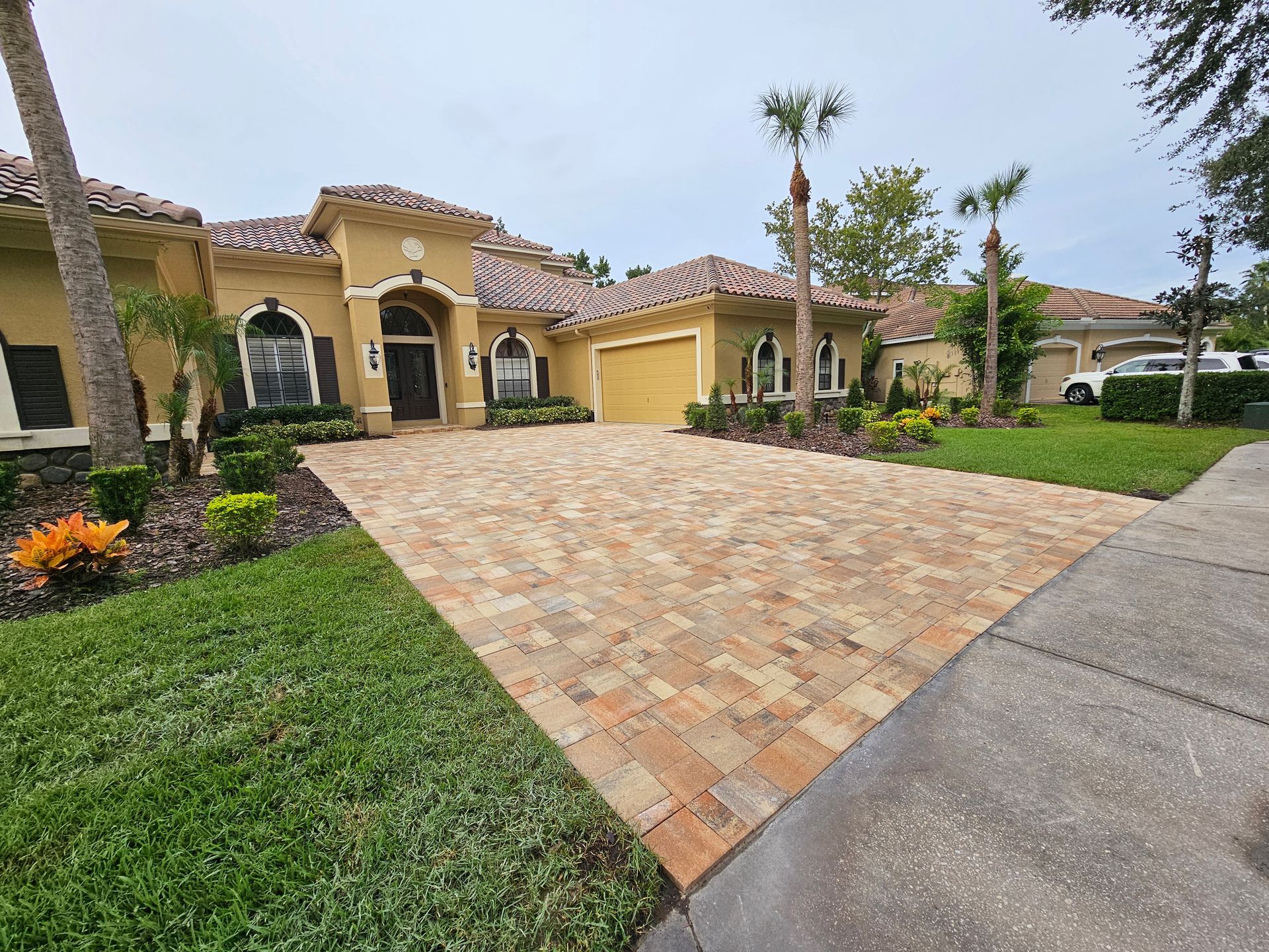 House with brick driveway, yellow exterior, palm trees, and green grass.