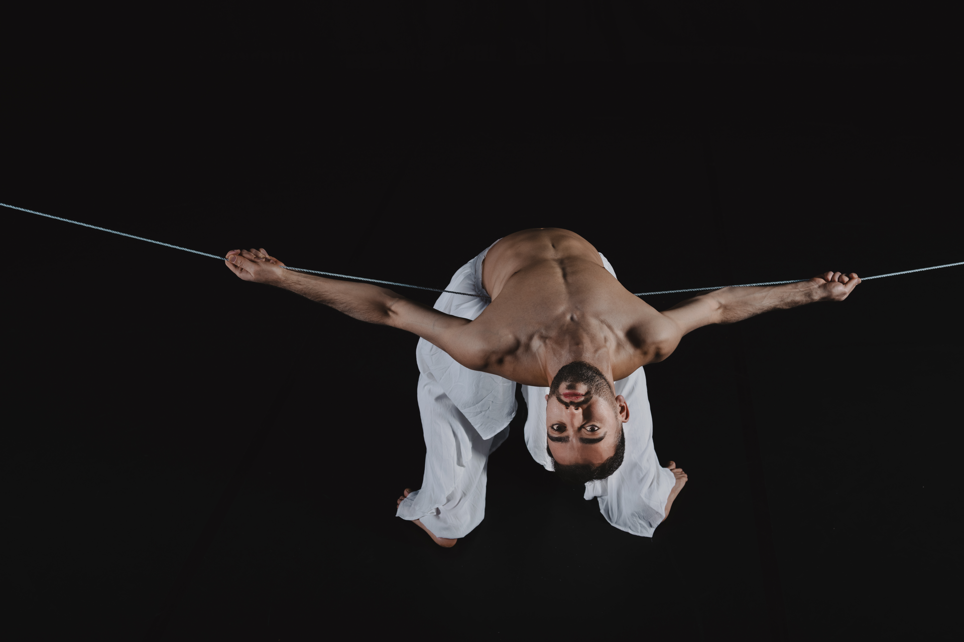 Een man in een witte broek hangt ondersteboven aan een strak gespannen touw, met uitgestrekte armen, tegen een zwarte achtergrond.