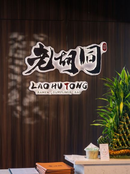 Restaurant sign on a dark wood wall reading “LAO HUI TONG,” with a plant and table in front