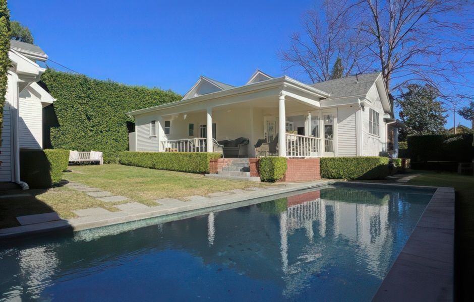 Backyard with a swimming pool, lawn, and white house with porch. Sunny day, blue sky.
