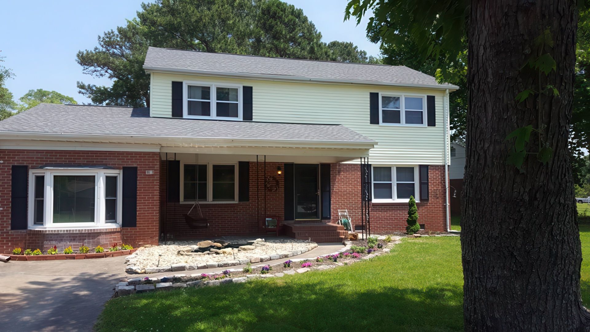 Two-story house with brick base and light siding. Black shutters. Green lawn and a large tree in the foreground.
