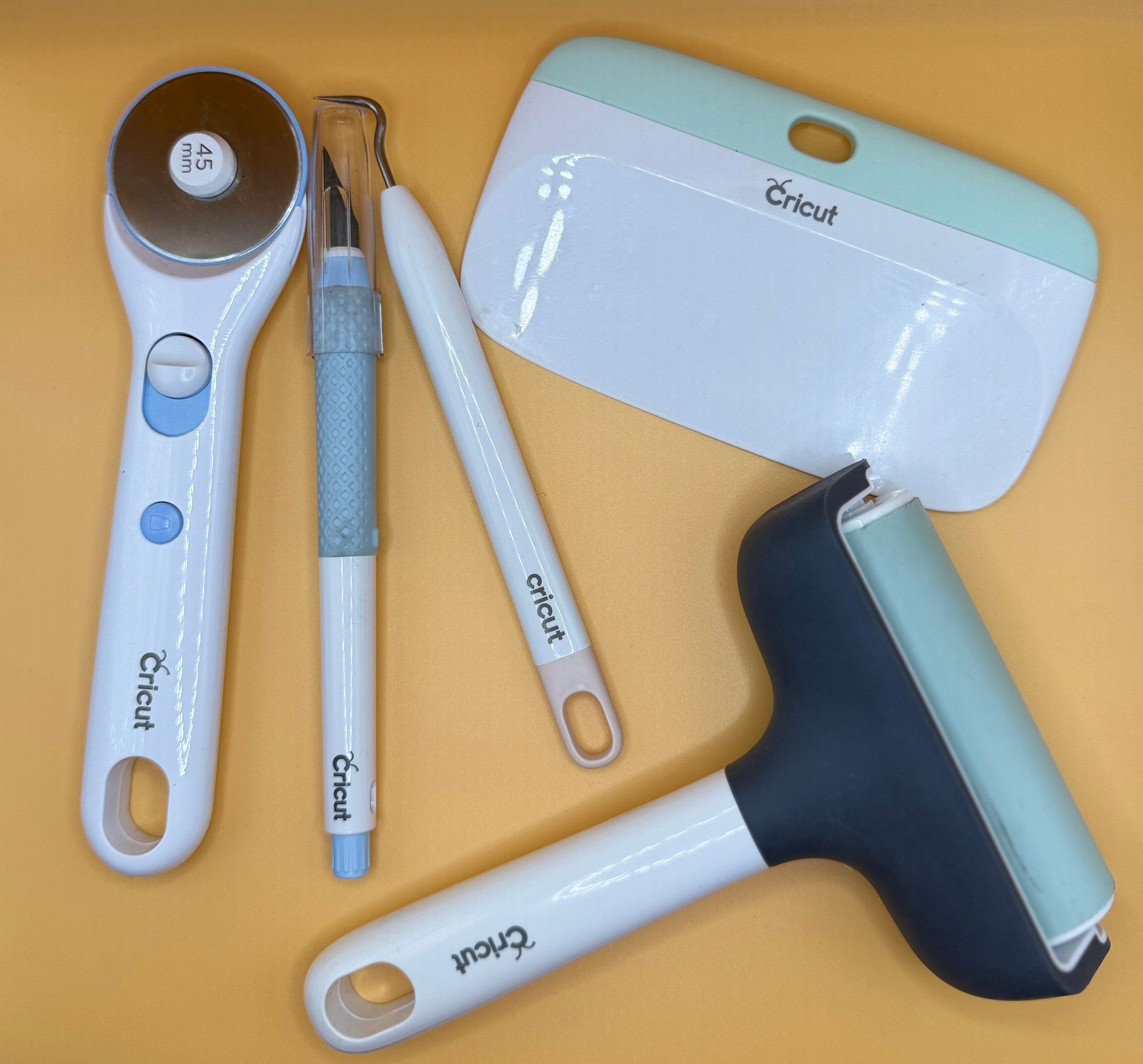 Cricut crafting tools: rotary cutter, weeding tool, scraper, burnisher, and lint roller on a yellow surface.