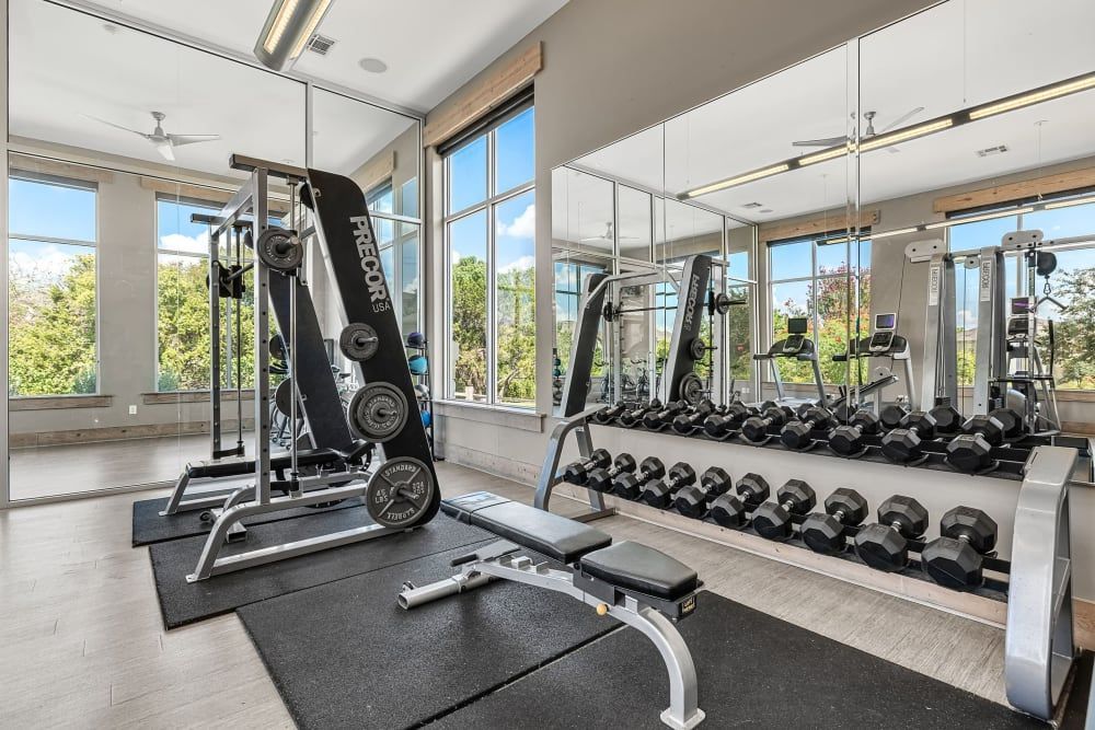 Interior of a bright gym with large windows on a sunny day, featuring a row of dumbbells on a rack, weight machines including a leg press and elliptical trainers, and an adjustable workout bench on a protective floor mat at Marquis at Canyon Ridge in Austin, TX.