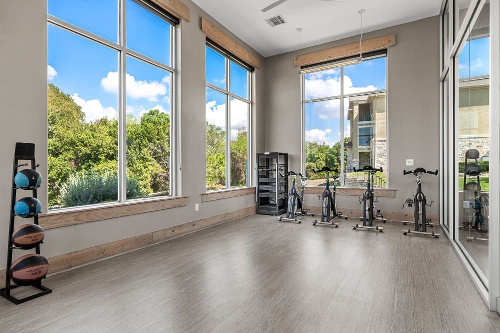Spacious, sunlit indoor gym with large windows overlooking greenery, equipped with stationary bicycles and a rack of medicine balls at Marquis at Canyon Ridge in Austin, TX.
