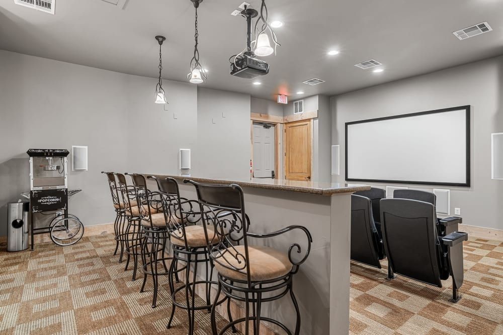 Modern home theater room with a bar counter featuring high stools, a vintage-style popcorn machine to the left, and three cinema-like seats facing a large projection screen. The room is well-lit with a mix of pendant lights and recessed ceiling lights, and the floors are patterned carpet. There's an exit door to the right of the screen at Marquis at Canyon Ridge in Austin, TX.