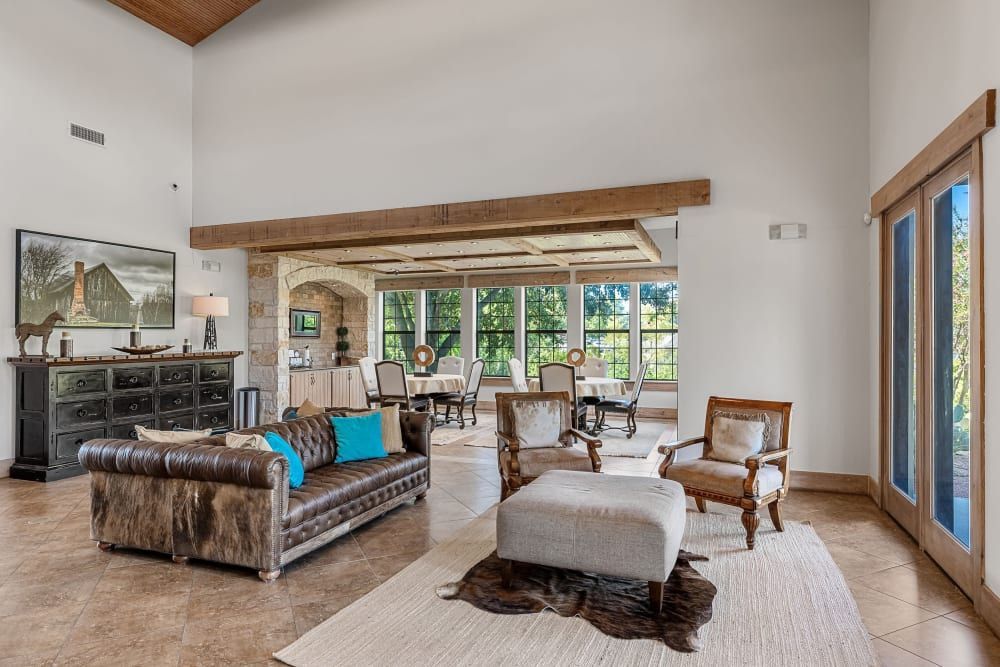 Spacious living area with high ceilings featuring exposed wooden beams, a large leather sofa with blue cushions, and two vintage wooden armchairs with white upholstery. In the background, a home office setup is visible behind an elegant stone archway, and natural light streams in through large windows and glass doors leading to the outdoors at Marquis at Canyon Ridge in Austin, TX.